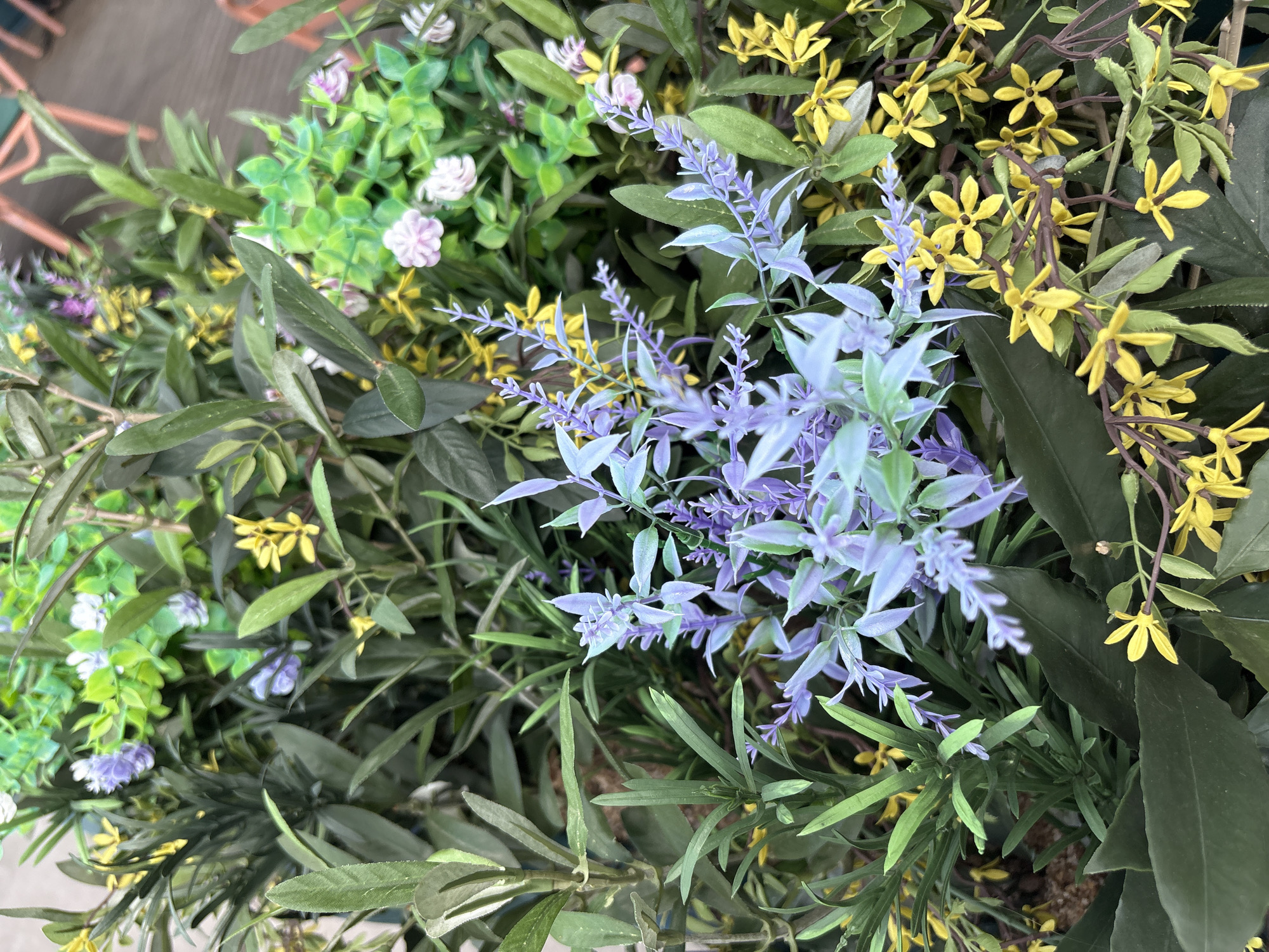 This is a close-up, overhead shot of a densely packed arrangement of artificial flowers and greenery. The subjects are the plants themselves, with a variety of colors and textures. There are prominent stalks of light purple flowers, resembling lavender, with delicate, clustered blooms. Interspersed are clusters of small, yellow flowers with five petals each, appearing like a delicate vine. Various shades of green foliage fill the spaces between the flowers, ranging from broad, dark green leaves to finer, lighter green stems and leaves. Small, white, clustered flowers are also visible, adding another layer of detail. The overall impression is of a vibrant and varied floral display.
The scene appears to be an outdoor setting, possibly a garden or a decorative arrangement at an event, given the fullness and artificial nature of the plants. There are no people or animals visible. The lighting suggests it could be daytime, with diffused light that doesn't create harsh shadows, hinting at possibly overcast weather or a shaded location. No text is visible in the image. The focus is entirely on the intricate details of the plant arrangement, showcasing the interplay of colors, shapes, and the textures of the artificial elements.