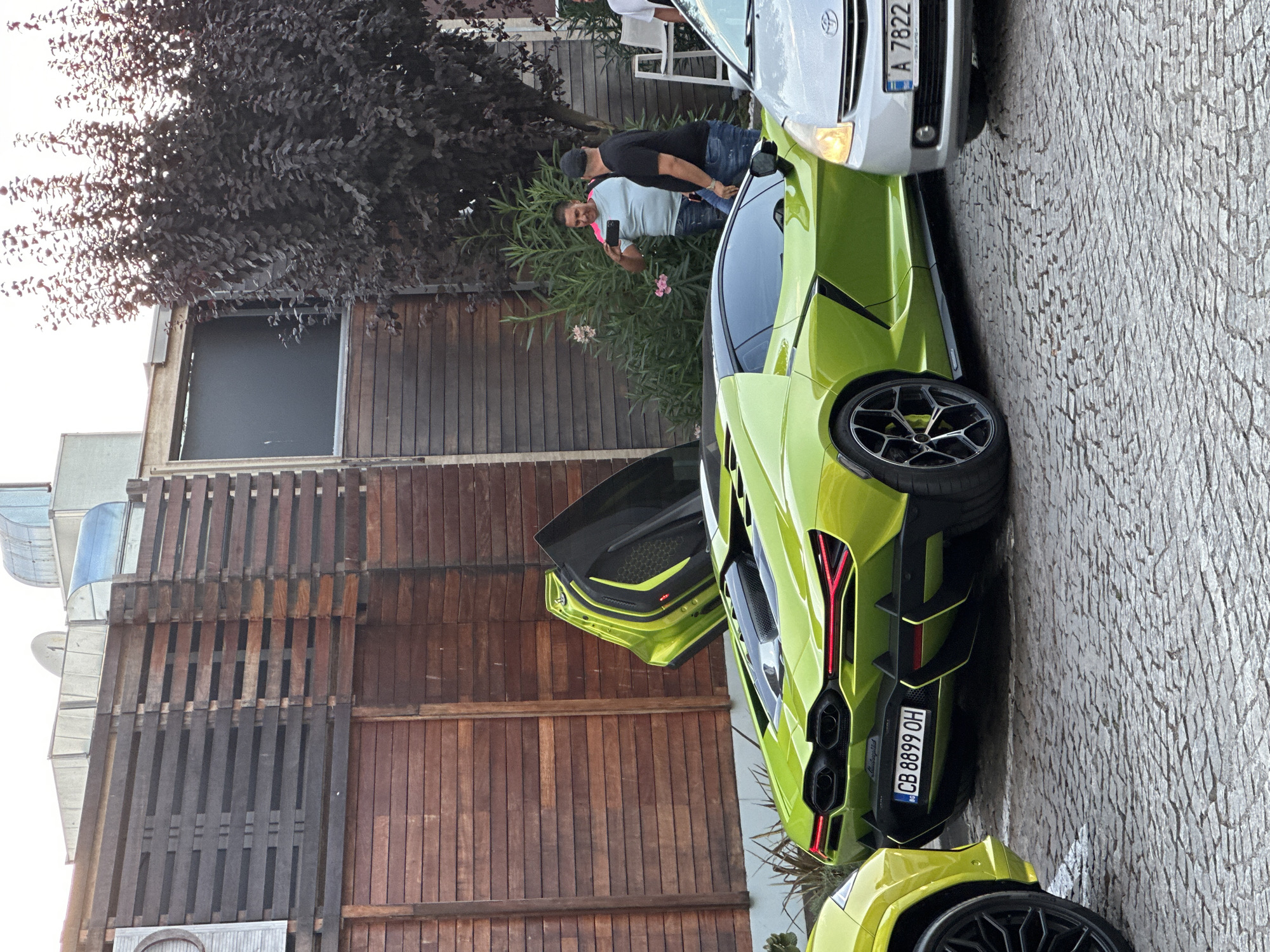 In Sozopol, Bulgaria, a lime green Lamborghini is parked on a cobblestone street, its doors open, showcasing its interior and exotic design. The car's license plate reads "CB 8899 OH." Adjacent to it, a silver Toyota Prius with license plate "A 7822" is also parked. Two men are standing near the cars; one, wearing a light blue t-shirt with pink trim and jeans, is holding a smartphone, seemingly taking a photo or video. The other man, in a black t-shirt and jeans, stands next to him. Behind the cars, a wooden fence with horizontal planks borders a building with visible architectural elements such as industrial-looking vents and a satellite dish. Lush green foliage and a dark-leaved tree contribute to the outdoor setting. The scene appears to be a casual gathering or showcase of vehicles.