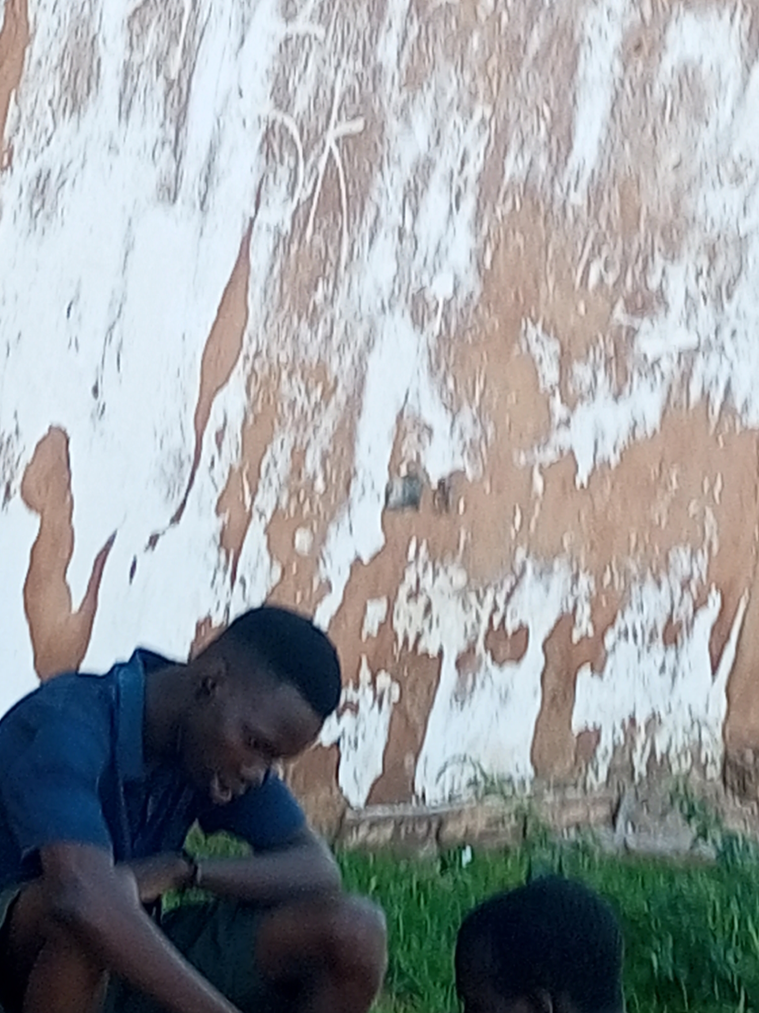 The image shows two young Black males in an outdoor setting, with a textured, peeling wall in the background and green grass in the foreground. The male on the left is wearing a blue short-sleeved collared shirt and dark shorts, and is crouched down with his head lowered, appearing to be speaking. He has a watch on his left wrist. The other male, on the right, is partially visible and only his head and shoulders are in frame, with his head bowed. The wall behind them is rough, with patches of white paint flaking off to reveal a brown, earthy surface. There are faint white markings on the wall, possibly graffiti or natural patterns. The overall scene appears to be in a somewhat dilapidated urban environment. The location is identified as Tempure, Nigeria.