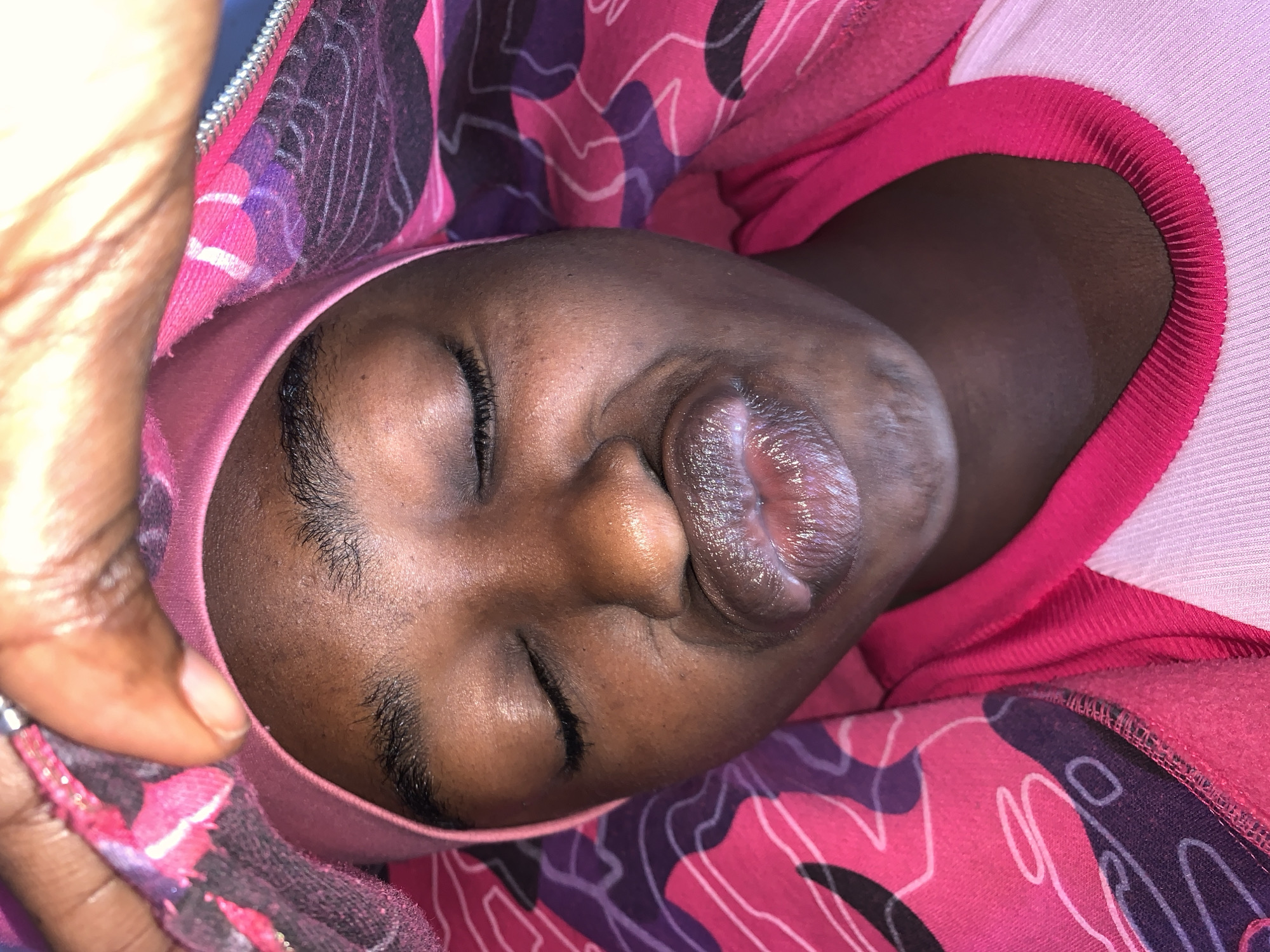 A close-up photograph depicts a Black individual, possibly an infant, asleep. The individual's eyes are closed, and their lips are slightly parted, exhibiting a moist appearance. They are wearing a pink hooded garment with a floral or abstract pattern in shades of pink, purple, and white. A human hand with tan skin is partially visible in the upper left corner, holding the edge of the hood.
There is no information that suggests this image was taken in Rafin Par, Nigeria.