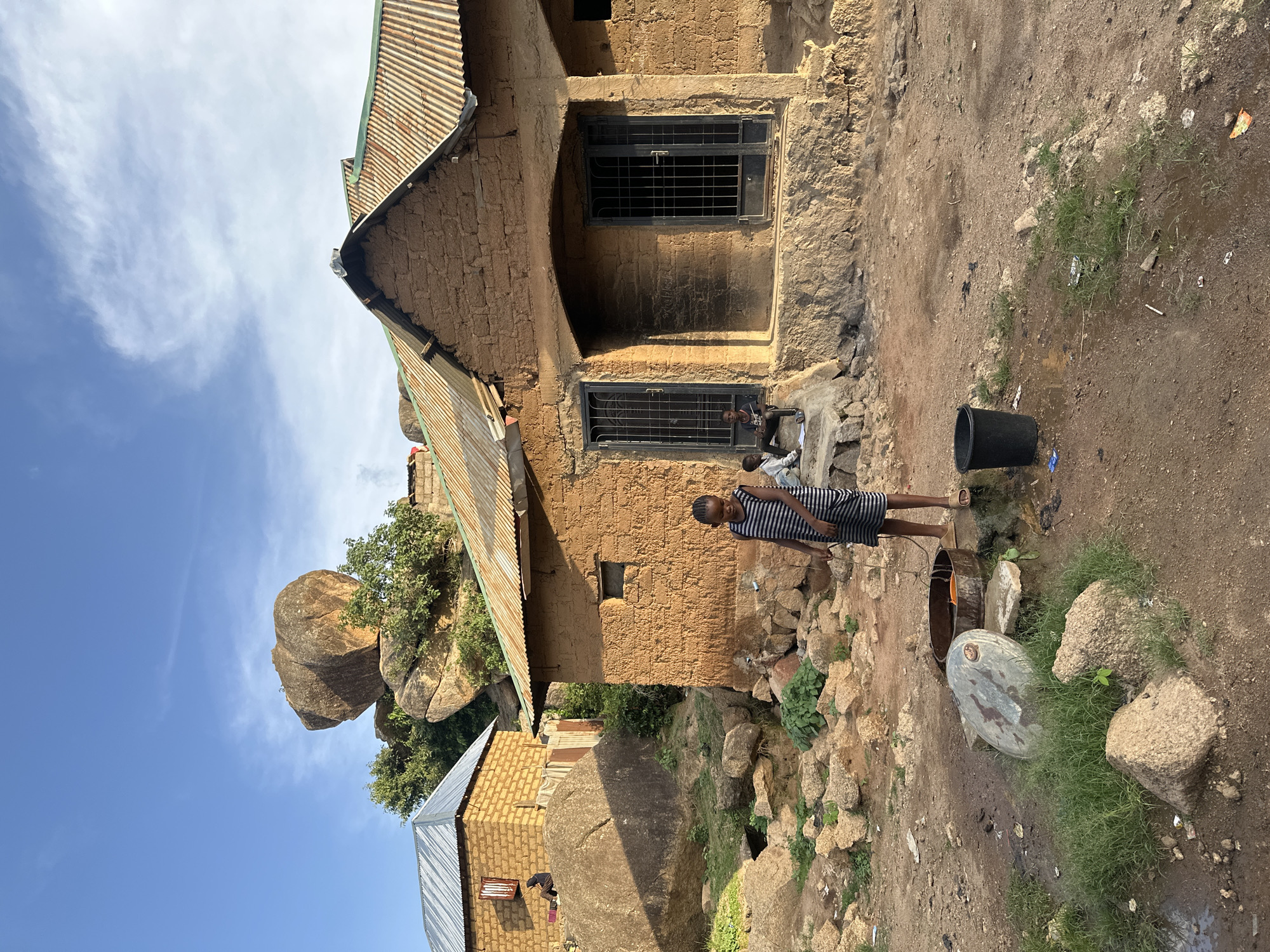 This daytime image, set in Jos, Nigeria, depicts a candid scene centered around simple, earth-toned buildings with corrugated metal roofs. The landscape is characterized by its rocky terrain, with large, weathered granite boulders (kopjes) dominating the background under a bright blue sky dotted with scattered white clouds.
In the foreground, a young person, possibly a child or teenager, stands barefoot on the dirt ground. They are dressed in a striped top and dark shorts or a skirt, appearing to observe something towards the left side of the frame. Nearby, a black plastic bucket sits on the dusty earth, alongside a larger, metallic basin or cut-open drum, which likely serves a practical purpose such as water collection or washing.
Closer to the main building, a smaller child is partially visible, sitting discreetly near the base of a window. The principal structure is built with robust mud-brick walls and features two windows secured with dark metal grates. The ground surrounding the buildings is a mix of dirt and scattered small rocks, interspersed with sparse patches of green grass and some visible debris. A second, smaller building of similar construction is partially seen on the far left.
The scene captures an everyday moment in a rural or semi-urban environment, highlighting architecture that blends with the rugged, rocky topography of the Jos Plateau. No discernible text is visible within the image.