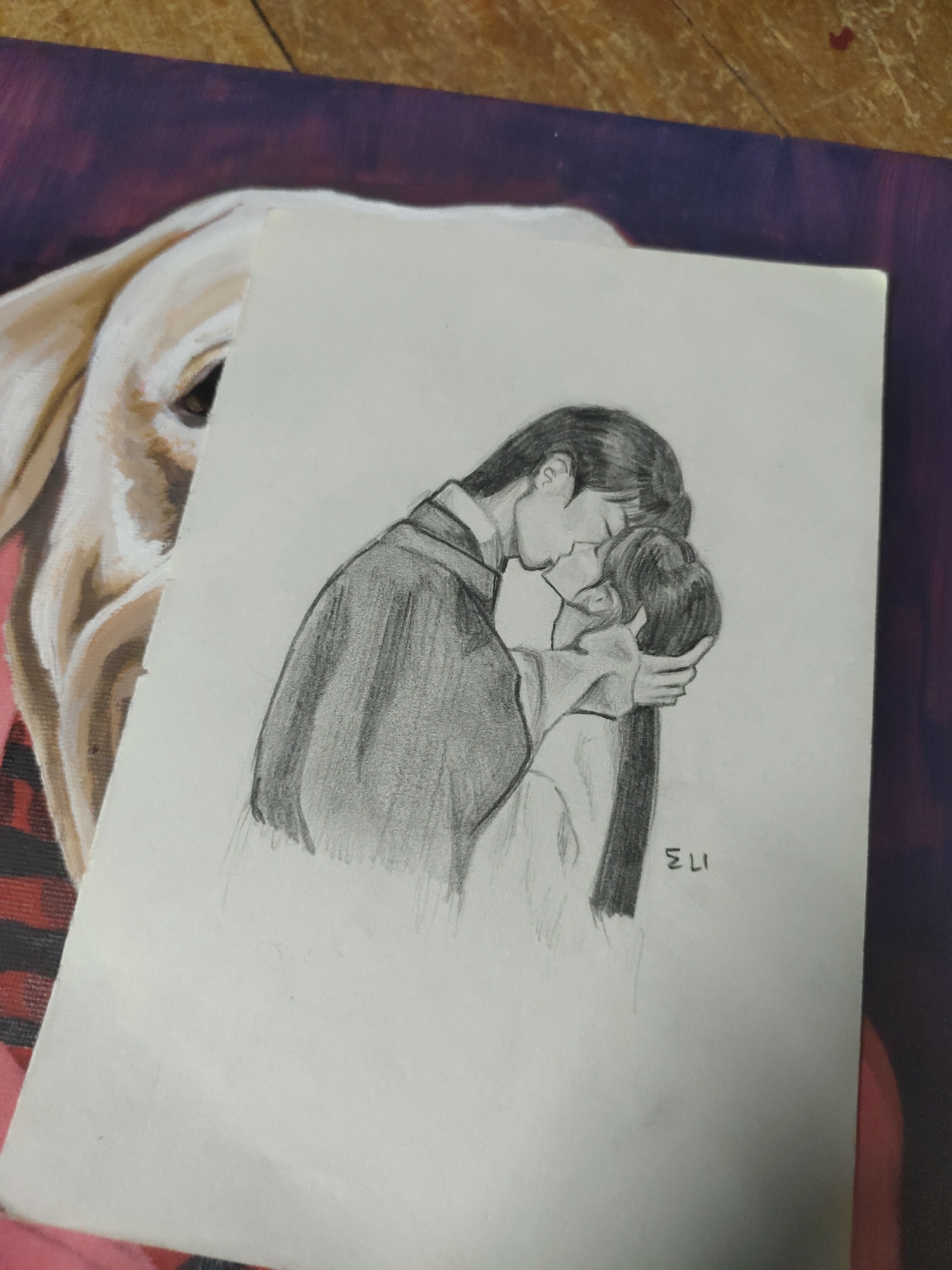 An overhead shot depicts a graphite pencil sketch on white paper, resting atop a larger, partially visible canvas painting. The sketch features two stylized figures, a male and a female, in a romantic embrace, their faces touching in a kiss. The male figure, positioned on the left, wears a dark, traditional East Asian-style robe with wide sleeves, and his left hand gently cradles the back of the female figure's head. The female figure, on the right, has dark hair styled back and wears a lighter garment. The bottom right of the sketch contains the handwritten signature "ELI 173". Beneath the sketch, the underlying artwork reveals the partial portrait of a cream-colored dog, likely a Labrador retriever, with one eye visible against a background of purple and red hues. This scene is situated on a wooden surface, displaying a distinct grain pattern, within a residential setting in Davao City, Philippines.