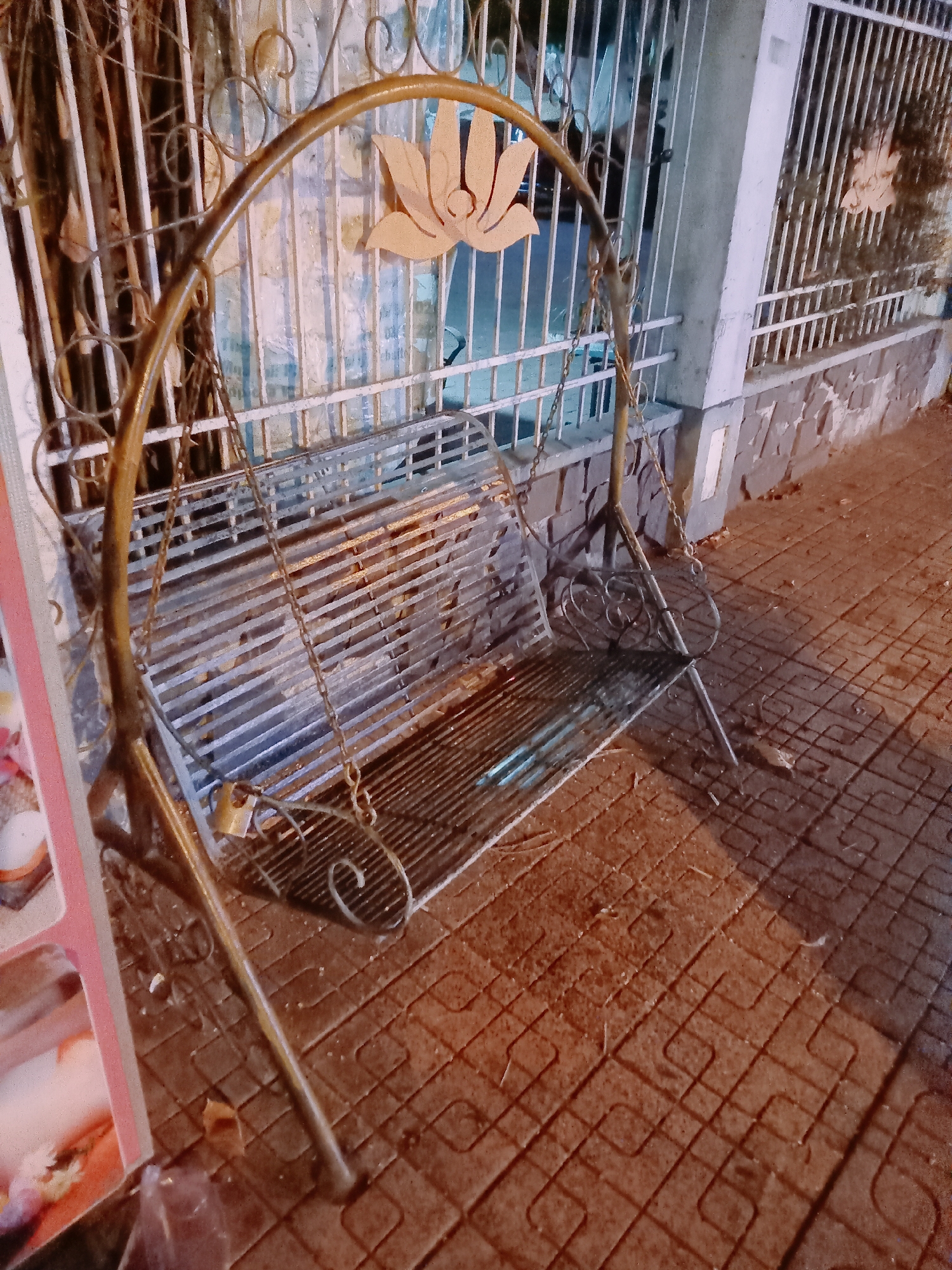 A metal swing bench sits on a brick-paved area in Nha Trang Ward, Vietnam. The bench has a decorative metal back with a lotus design. It's suspended by chains and secured with a padlock. Behind the bench is a wall with a barred fence. The lighting suggests it is nighttime.