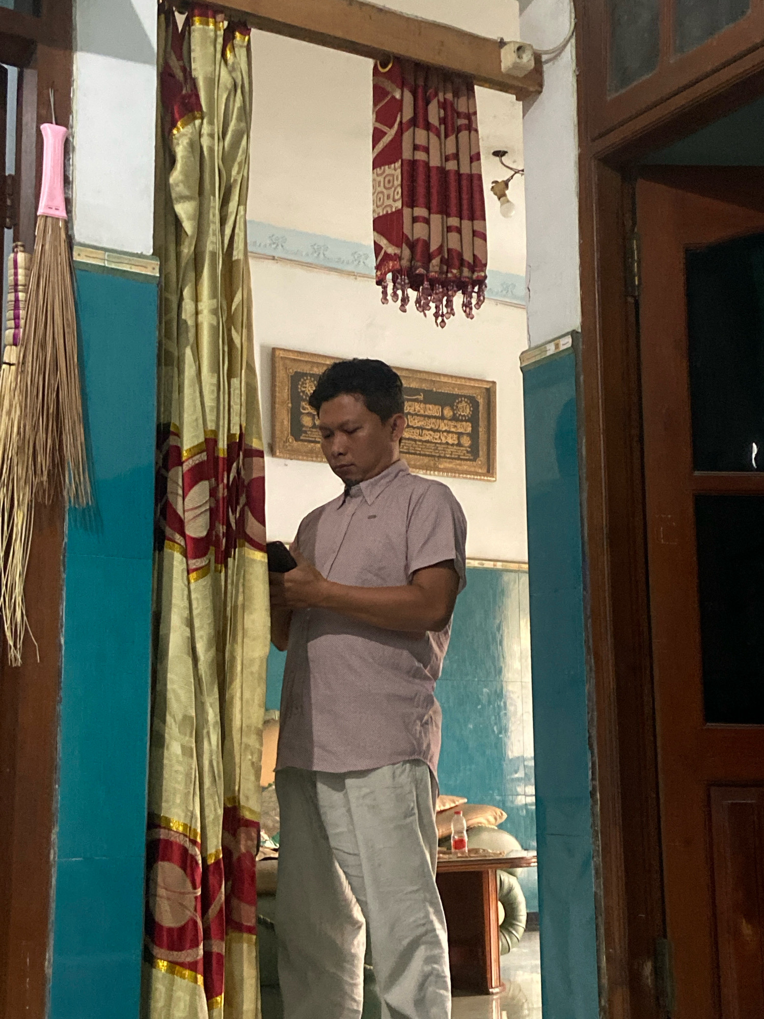 A man in a light purple button-down shirt and light grey pants stands in what appears to be the interior of a home in Gelam, Indonesia. He is holding a dark-colored smartphone in his hands and looking down at it. Behind him, a large curtain with a red and gold circular pattern hangs from a wooden beam. To his left, a broom with a pink handle is leaning against a turquoise wall. Further in the background, a framed piece of artwork with Arabic script hangs on the wall, and a table with pillows and a water bottle is visible. A wooden door frame is to the right of the frame.