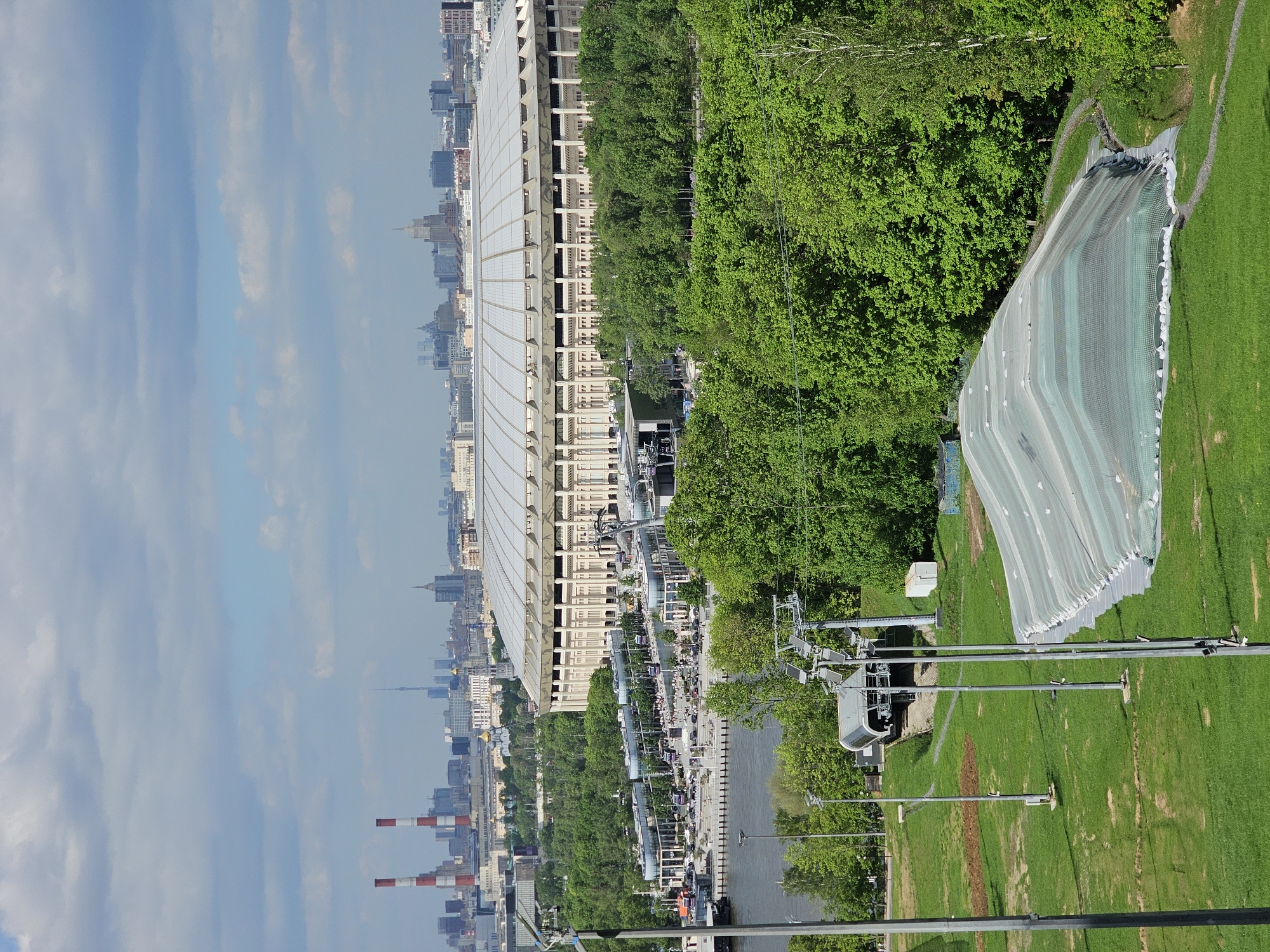 The image captures a view in Moscow, Russia, likely in the area of Luzhniki Stadium and the Sparrow Hills. In the foreground, there is a ski jump covered with a light-colored mesh, surrounded by green grass. An aerial tramway with gondolas is visible, running parallel to the ski jump. Moving towards the left of the image, trees line the edge of a river or body of water, along which there's a pedestrian walkway populated with people. Adjacent to this, there is a large building, possibly the Luzhniki Stadium, with a distinctive roof structure. In the background, a cityscape extends into the distance under a cloudy sky. There are visible smokestacks and various other buildings in the urban landscape.