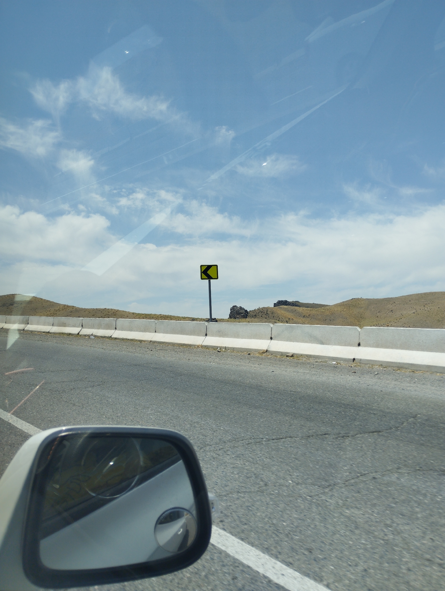 The image depicts a road scene in Jizzakh, Uzbekistan. A black and yellow road sign indicating a sharp left turn is visible on the right side of the road, mounted on a pole. The road is paved with asphalt and has white lane markings. A concrete barrier runs along the side of the road, separating it from a dry, hilly landscape with sparse vegetation. The sky is blue with scattered white clouds. The reflection in the car's side mirror shows the interior of the car. The overall impression is a view from a moving vehicle on a highway in a semi-arid region.