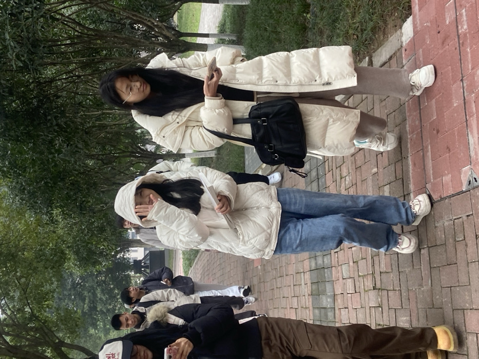A group of Asian people stand outdoors on a brick walkway. Several wear winter coats. One woman holds a phone. Another has her hood up. Trees are visible in the background.