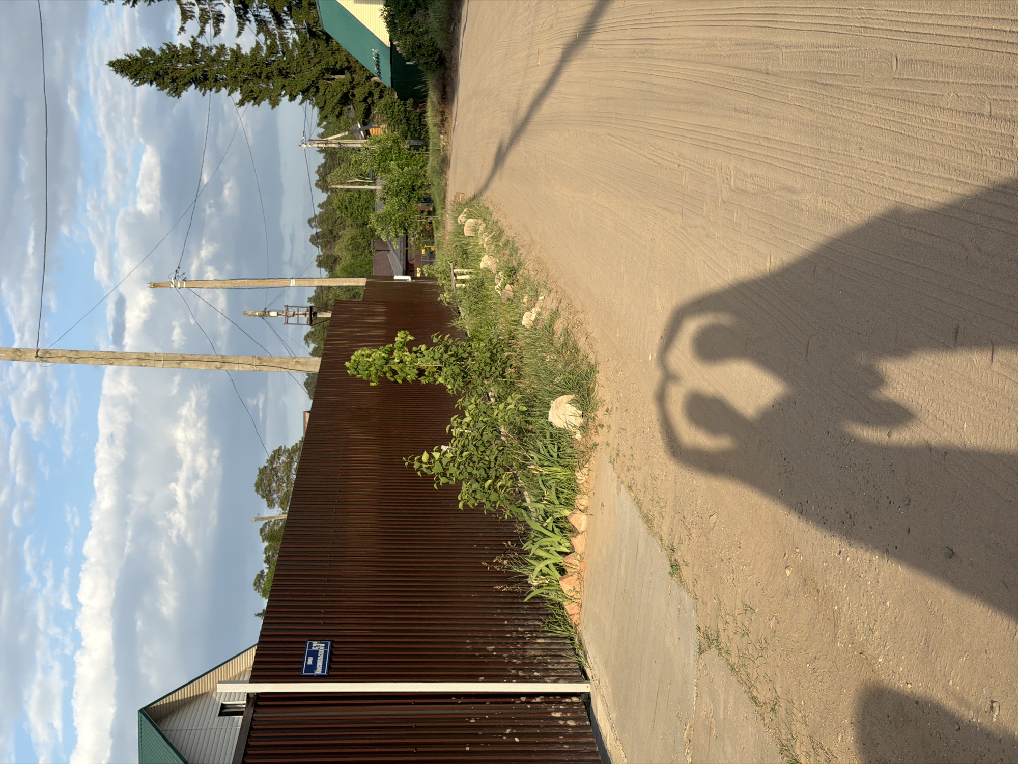 The image depicts a rural street scene in Chaikovsky, Russia. The view is along a dirt road bordered by a brown corrugated metal fence on the left and vegetation along the edge of the road. A portion of a house with a green roof is visible behind the fence. Power lines are strung between wooden poles that run parallel to the road. The sky is visible with clouds. The shadow of a person with an arm raised is cast on the road surface, indicating the presence of a person taking the picture, and an address sign in Cyrillic is mounted on the fence.