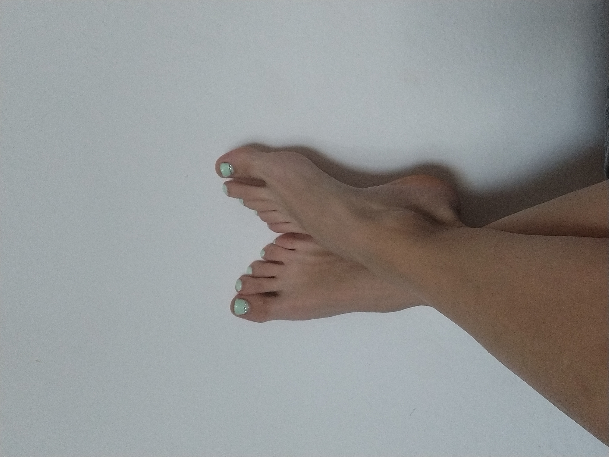 This is a close-up, waist-up photograph of a person's bare feet crossed against a plain, light-colored wall, likely indoors. The feet are positioned in the lower half of the frame, with the soles facing slightly upwards. Both feet are neatly presented, with light green nail polish applied to the toenails, some adorned with small, clear rhinestones. The lighting is soft and diffused, creating subtle shadows that highlight the contours of the feet and legs. There is no visible interaction or action; the focus is solely on the aesthetic presentation of the feet. The background is a simple, textured wall with no discernible features, suggesting a domestic or studio setting. There is no information in the image to determine the location in Dobev, Czechia.
