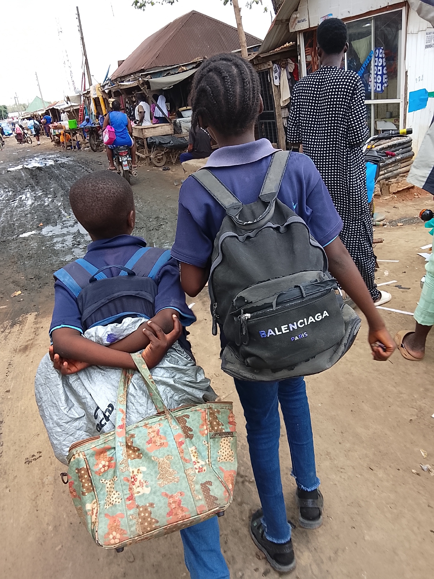 Two children walk down a dusty street. One child wears a blue shirt, blue jeans, black shoes, and carries a large, blue floral-patterned bag and a gray plastic bag. The other child wears a blue shirt, blue jeans, black shoes, and a gray backpack labeled "BALENCIAGA PARIS". In the background are shops and stalls, with people milling about. The street is dirty and muddy in places. The location is Abuja, Nigeria.