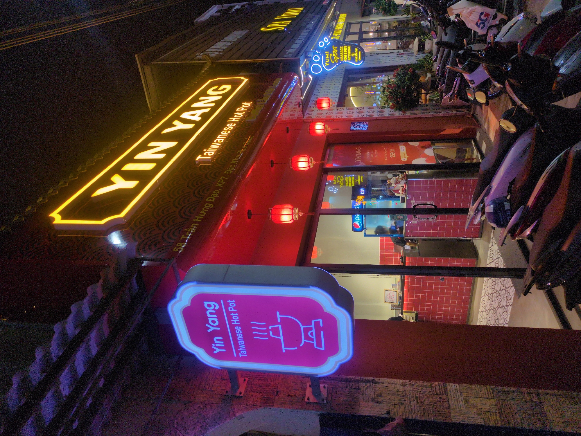 The image depicts the exterior of the "Yin Yang Taiwanese Hot Pot" restaurant located at 58 Trần Hưng Đạo, KP7, Đặc Khu Phí mộc, Phú Quốc, Vietnam, at night. A yellow neon sign displaying "YIN YANG" is above the building, with "Taiwanese Hot Pot" written below it. A smaller, pink neon sign with an illustration of a hot pot also displays "Yin Yang Taiwanese Hot Pot." The building is adorned with red lanterns. A "YUMI Spa" sign is visible above the restaurant. Parked motorbikes are in front of the building. Through the restaurant's windows, portions of the interior are visible, including red tiled walls and people are present.
