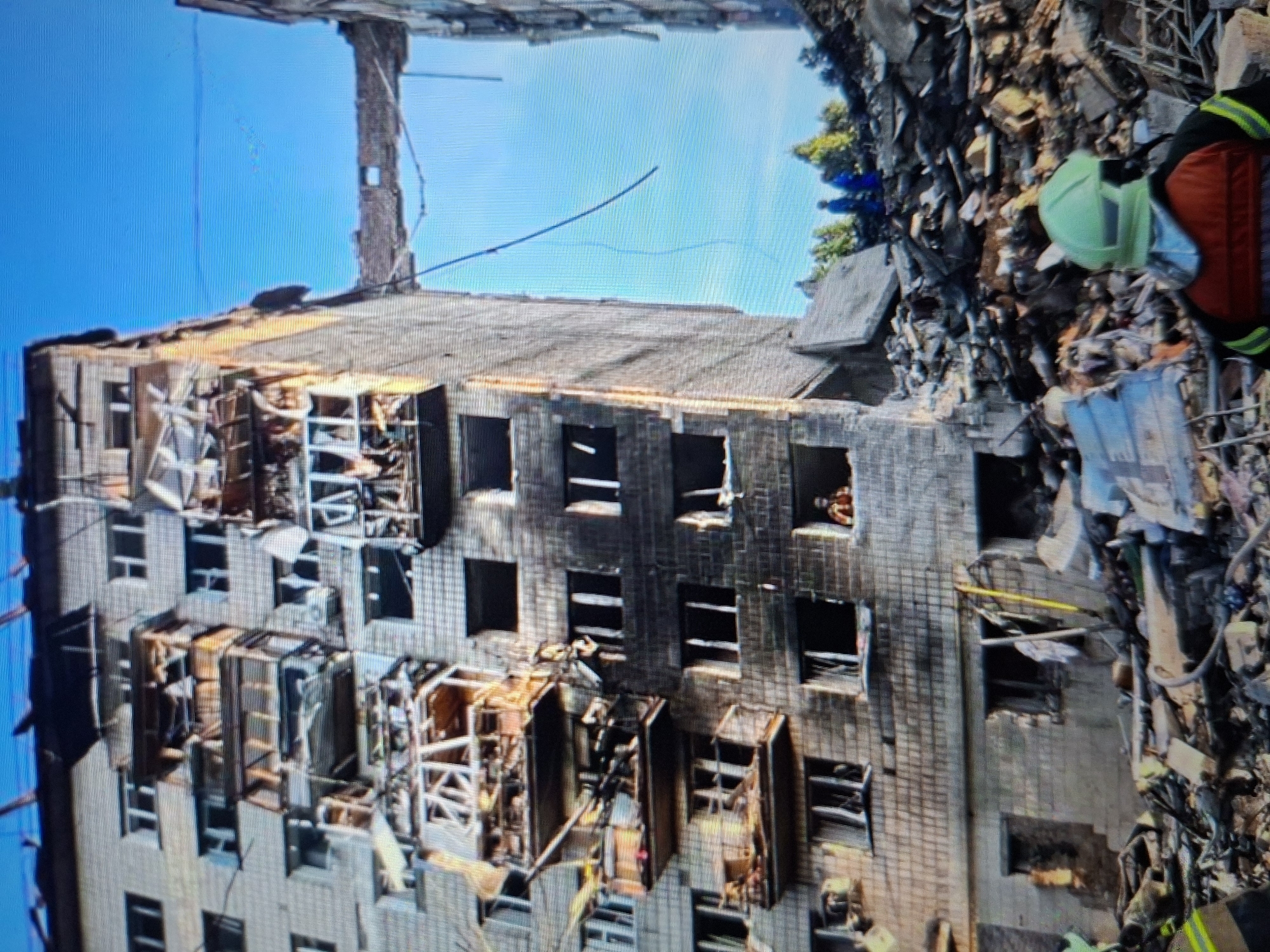 The image depicts the aftermath of an event that has caused significant damage to a multi-story building. The building, constructed of light-colored brick, shows extensive damage to its facade, with many windows broken or missing. Balconies on multiple floors have collapsed or are severely damaged, with exposed scaffolding and debris. The upper floors appear to have suffered the most, with large sections of the building missing and debris piled high to the right of the structure.
In the debris to the right, several individuals can be seen, likely involved in rescue or recovery efforts. One person is wearing a bright orange and red vest, suggesting they are a rescuer. A green hard hat is also visible among the rubble, and another individual appears to be wearing a similar protective helmet and yellow safety vest. The presence of these individuals and the evident destruction indicate a disaster or emergency situation.
The sky in the background is a clear, bright blue, suggesting daytime and fair weather. The scene appears to be an urban environment, given the multi-story building. The overall impression is one of devastation and ongoing efforts to deal with the consequences of a destructive event. There is no visible text in the image.