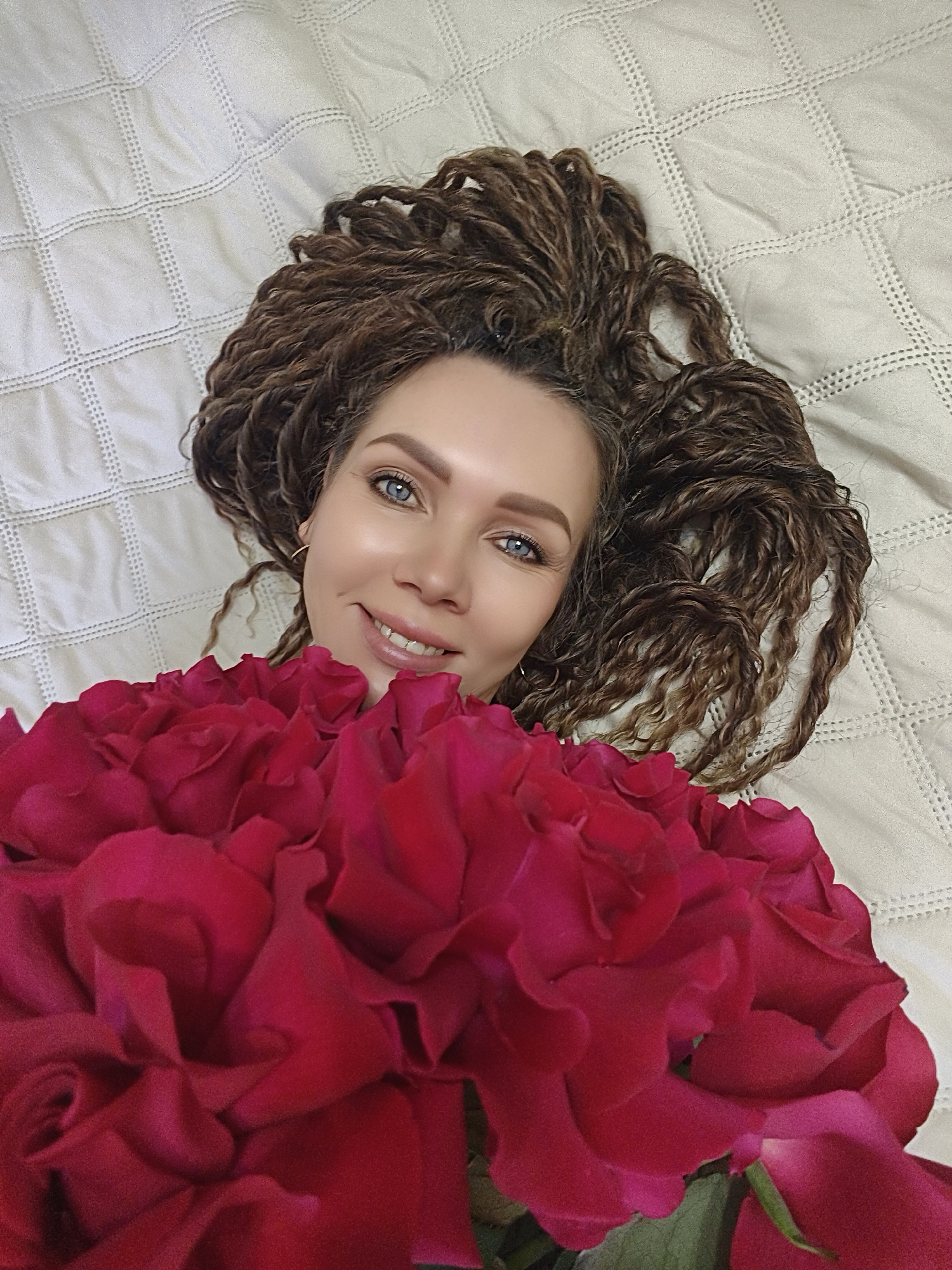 A woman with light skin and blue eyes, possibly in her late 20s to mid-30s, lies on a light-colored, quilted bedspread. She is smiling and holding a large bouquet of red roses close to her chest. Her brown, styled hair is spread out around her head on the bed. No other identifying information or location details are discernible.
