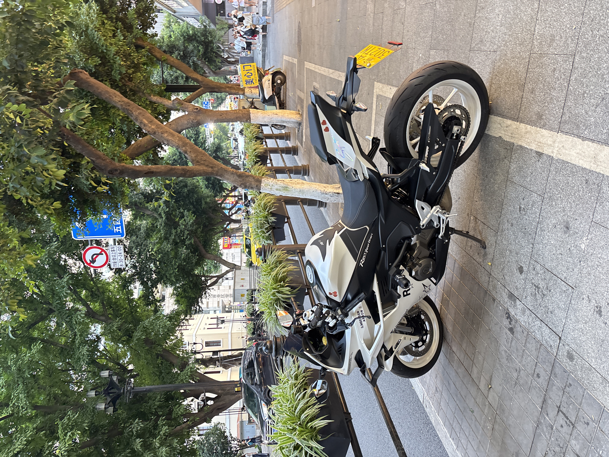 This is a medium shot, eye-level, full outdoor photograph taken in Yuzhong District, China. In the foreground, a white and black sport motorcycle is parked on a tiled sidewalk. The motorcycle is angled to the right, with its front wheel closest to the viewer. The motorcycle has a black windshield, silver handlebars, and various decals, including "R," "Rev-Your Heart," and two small red hearts. The rear license plate, which is yellow, is partially obscured by shadows. The motorcycle's kickstand is deployed, and its rear tire is large and black with white spokes on the rim.
Behind the motorcycle, a metal railing separates the sidewalk from a street. The street is lined with planters containing variegated foliage. Beyond the planters, a black car and a yellow taxi are visible on the street. Several people are walking on the sidewalk in the background, and a white and blue moped is parked further down the street. Traffic signs are present in the upper portion of the image, including a red circle with a left-pointing arrow, and a blue sign with Chinese characters. Mature trees with dense green foliage line the street and sidewalk, casting shadows. Buildings are visible in the background, with windows and architectural details. The overall lighting suggests it is late afternoon or early evening.