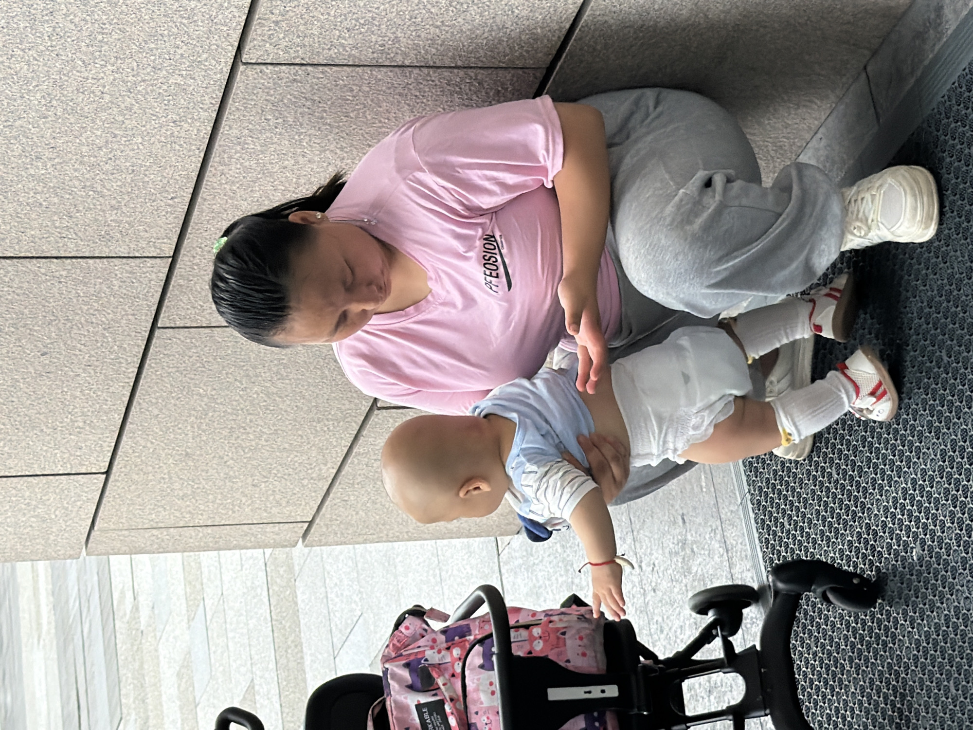 In Yuzhong District, China, a woman wearing a pink t-shirt and grey sweatpants crouches beside a stroller, holding a baby dressed in a blue and white striped shirt and a diaper. The baby, facing away from the viewer, is reaching out with its left hand, while its right hand is being held by the woman. The stroller, patterned with cats, is positioned to the left of the woman and baby. The scene takes place outdoors next to a stone wall.