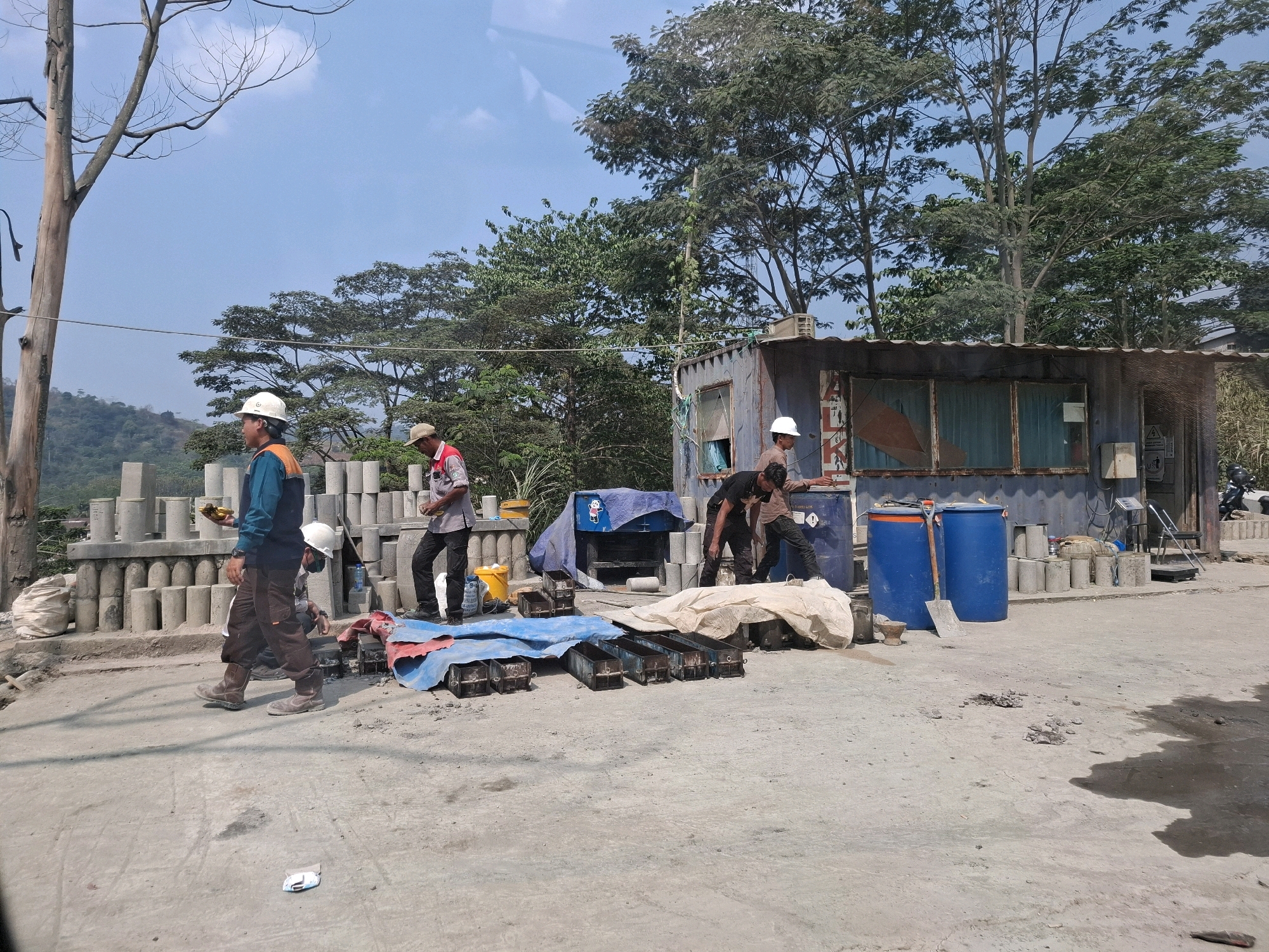 The image depicts a construction site in Cijangkar, Indonesia, under a clear blue sky. Several men in work attire, including hard hats, are present. In the foreground, a man wearing an orange and blue uniform with a white hard hat walks to the left, carrying a yellow object. Behind him, several cylindrical concrete forms are stacked. To the right, a group of men are gathered near a corrugated metal structure that appears to be a temporary office or storage unit. Two large blue barrels stand near this structure, one with a shovel leaning against it. Various construction materials, including tarps, metal molds, and bags of material, are scattered around the site. The environment is dusty and appears to be an outdoor industrial area with trees and hills visible in the background.