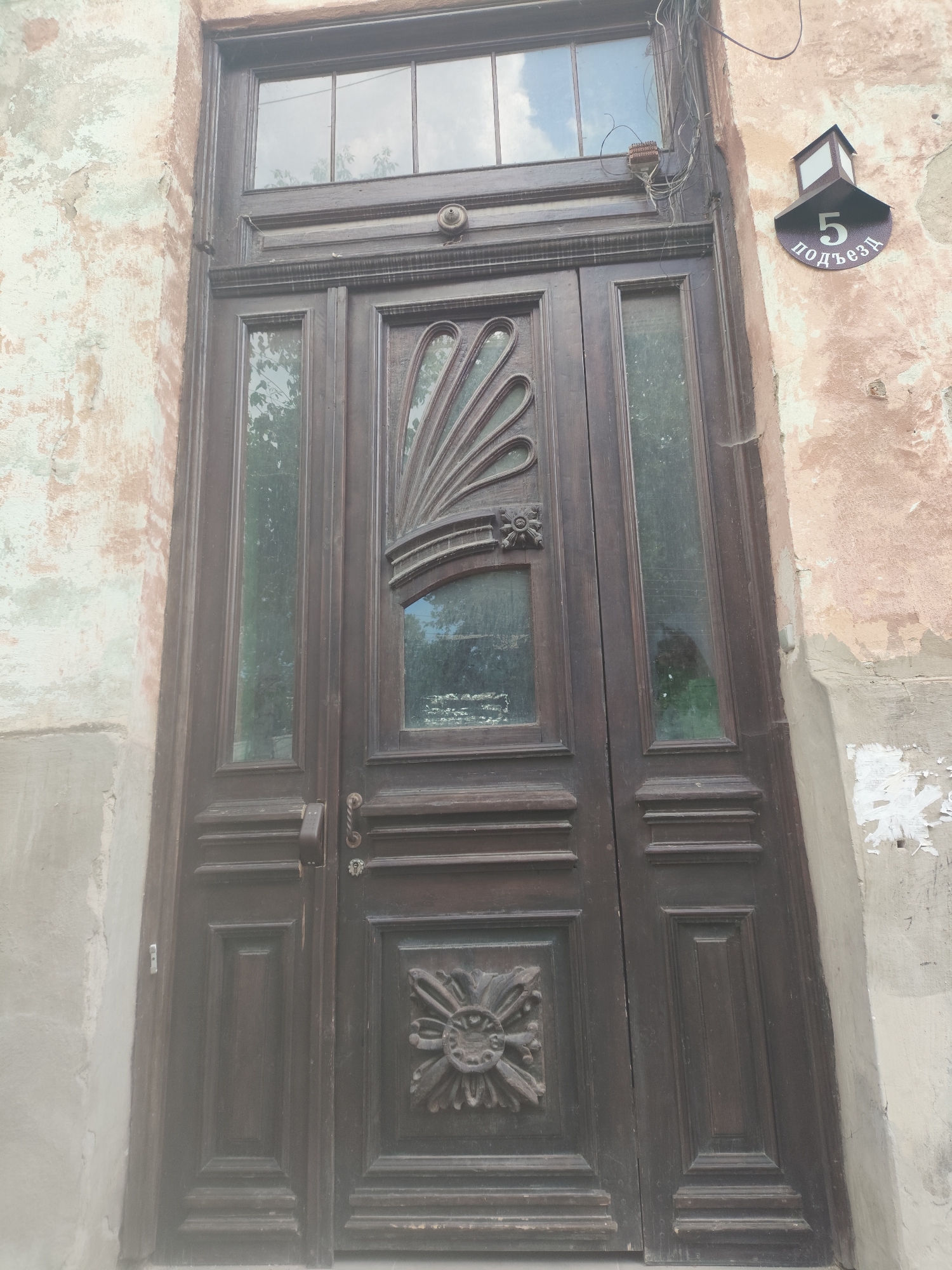 The image shows a dark wooden double door with glass panels, located in Simferopol, Ukraine. The door is adorned with decorative carvings. Above the door, there are windows with visible reflections of the sky and trees. To the right of the door, mounted on a pink, weathered wall, is a dark sign with the number "5" and Cyrillic text reading "ПОДЪЕЗД". Wires are visible near the top of the door and extending from the wall. The wall itself shows signs of peeling paint and age.