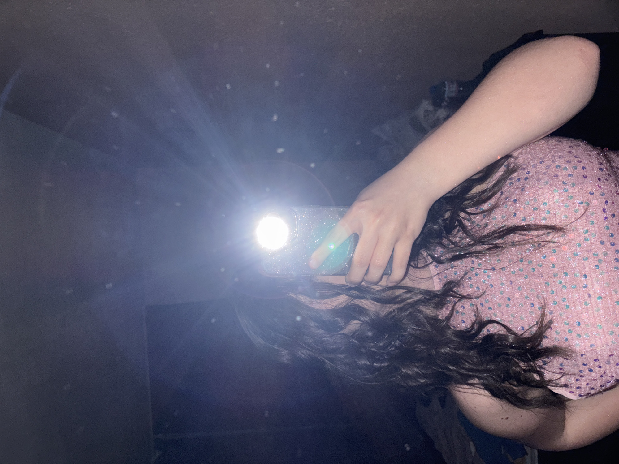 A person is taking a mirror selfie in a dimly lit room. The phone's flash is on, causing glare. The person is wearing a pink sequined top. The location is unknown and there is no indication of Garmen, Bulgaria.