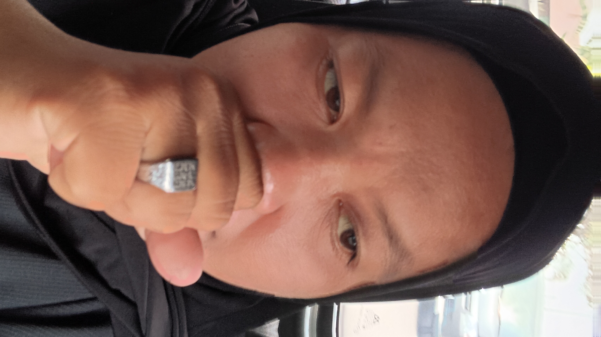 The image is a close-up portrait of a person wearing a black hijab, covering their head and neck. The person's face is partially obscured by their hand, which is covering their mouth and nose. The person is wearing a silver ring with a square top on their right ring finger. The background is out of focus but appears to be an outdoor environment, possibly a street scene with vehicles visible. The lighting suggests it is daytime. The person's skin tone is medium to dark brown. Their eyes are visible, and they appear to be looking slightly to the right of the camera.