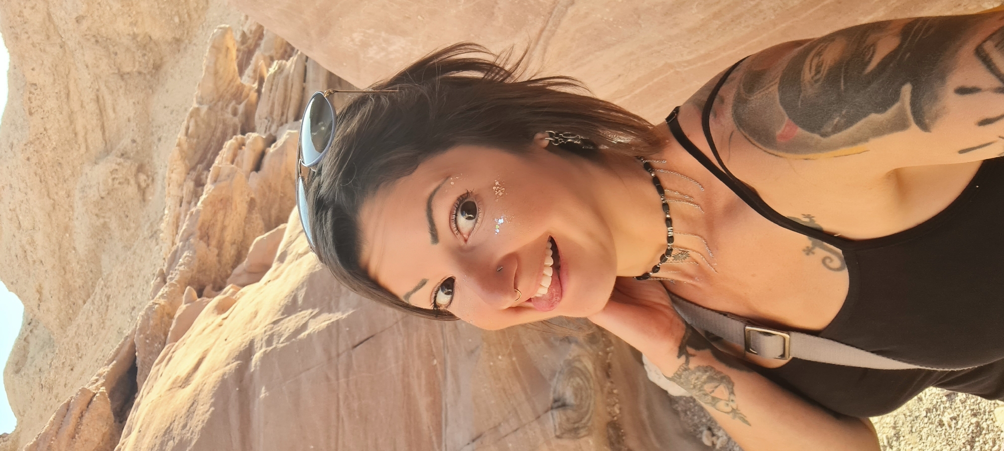 A woman with short brown hair, sunglasses perched on her head, and glitter around her eyes poses for a photograph against a backdrop of light brown rock formations. She has a nose ring, a necklace with dangling silver chains, and tattoos on her arms and chest. She is wearing a black tank top and a grey strap crosses her chest. The location is Hevel Eilot Regional Council, Israel.