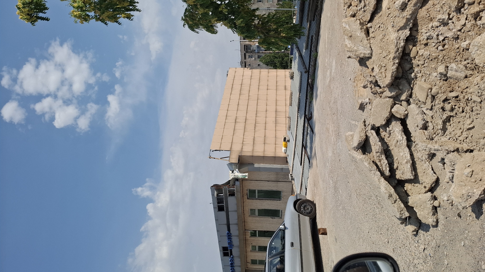 The image depicts a construction or demolition scene in Shymkent, Kazakhstan. In the foreground, a pile of rubble, consisting of broken concrete and dirt, is visible on a dusty ground. To the left, a silver sedan is parked on a paved surface, with a small wooden block near its front wheel. A side mirror of a car is also partially visible on the far left.
The background features a damaged building. The left section of the building has a gray facade with visible signage in Cyrillic letters that reads "СПОРТОВЫЙ ЦЕНТР" (Sports Center). This section appears to be partially collapsed or under renovation, with exposed structural elements and debris. Adjacent to it, on the right, is a large, light brown tiled wall. In front of these buildings, a corrugated metal roof structure is laid out, supported by metal beams.
Further in the background, a multi-story apartment building is visible, suggesting an urban setting. Green trees, including one with yellow flowers, are present on the right side of the image. The sky is blue with scattered white clouds. The overall impression is one of ongoing construction or repair work in a city environment.