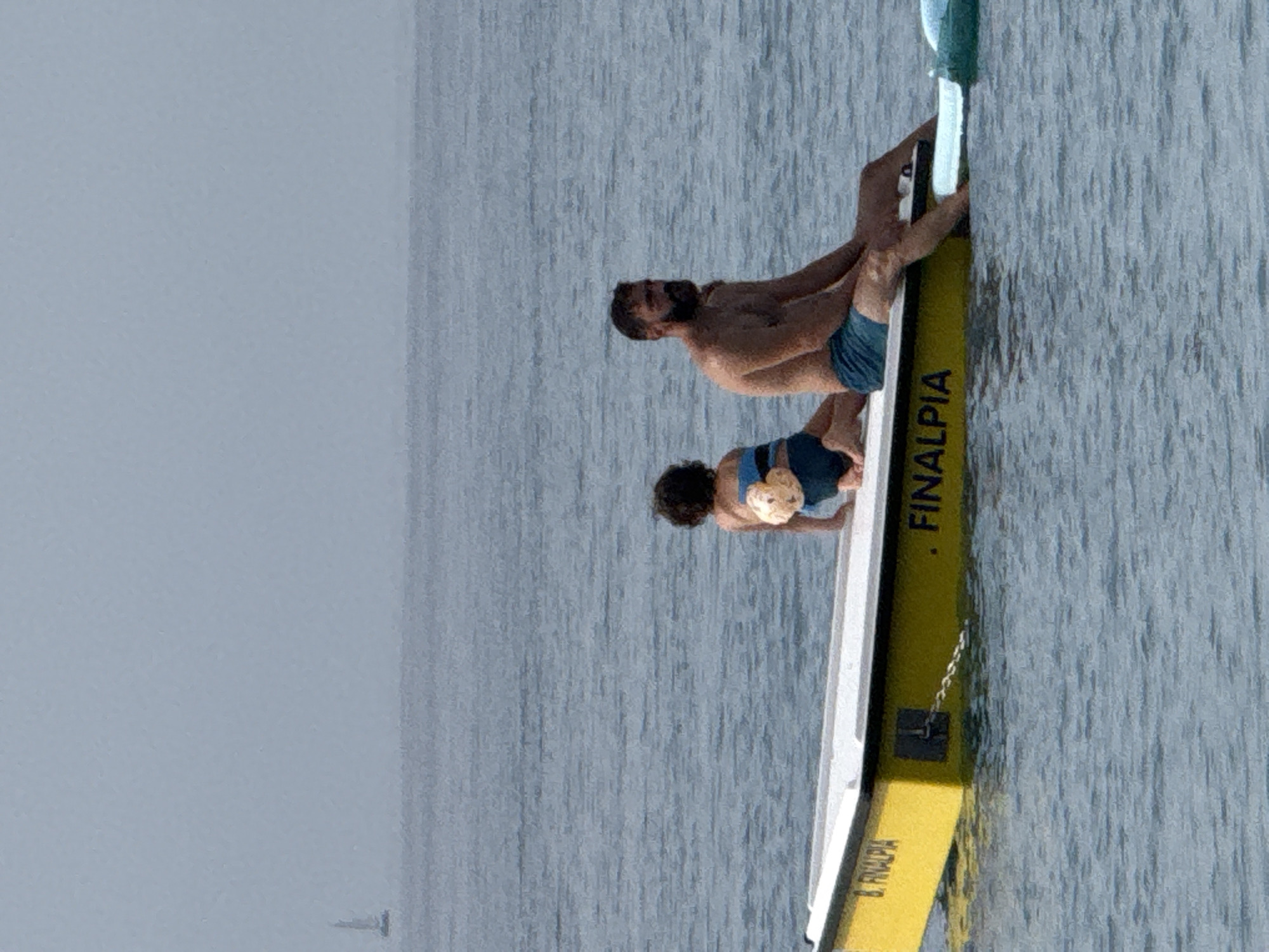 In Finale Ligure, Italy, a yellow and white boat floats on calm blue water. Two individuals are seated on the boat, facing away from the camera. A man with a beard, wearing blue swim trunks, sits on the right side of the boat. To his left, a child with curly hair, wearing blue swim trunks, is perched with their back to the viewer. The boat has the word "FINALPIA" written in black on its yellow side, and the letter "B." is visible on the left side. In the distance, a sailboat can be seen on the horizon under a hazy sky.