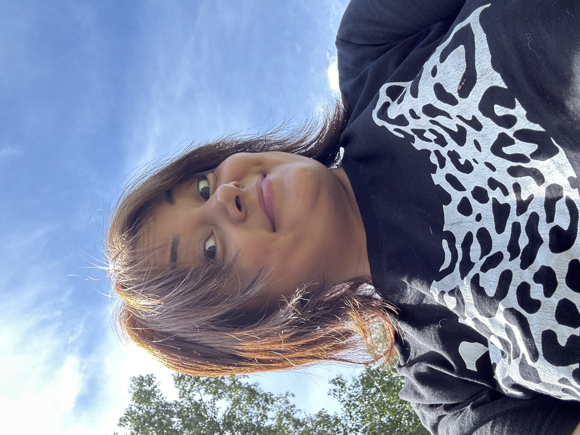 A low-angle, waist-up shot captures a woman outdoors against a blue sky with sparse clouds. She has medium-length, auburn hair that is slightly windswept, framing her face. She is looking directly at the camera with a playful, slightly surprised expression, her eyes wide and mouth curved into a smile. Her skin appears tanned. She is wearing a black t-shirt with a large, white leopard print design across the chest. To the left of the frame, the blurred greenery of trees is visible. The lighting suggests it is daytime, with sunlight illuminating her face from above.