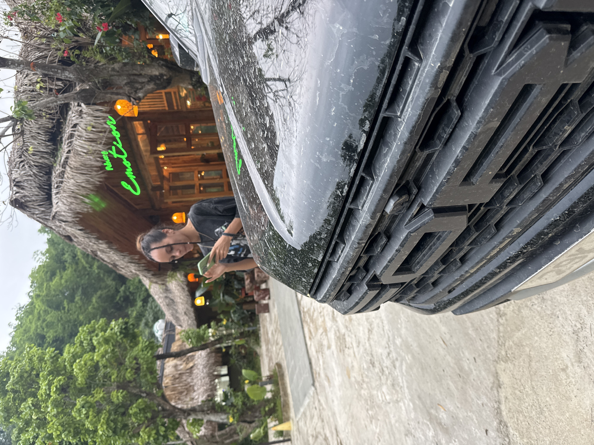 A person, appearing to be a young woman with dark hair pulled back, is standing outdoors and looking at a smartphone. She is wearing a dark t-shirt. In the background, a building with a thatched roof is visible, with glowing orange lights and green neon signage that reads "Music Emotion." The building is surrounded by trees and lush vegetation. In the foreground, the hood and grill of a black vehicle are prominently displayed, appearing to be slightly dirty and wet. The overall scene suggests an outdoor setting, possibly a restaurant or cafe, in a natural environment.