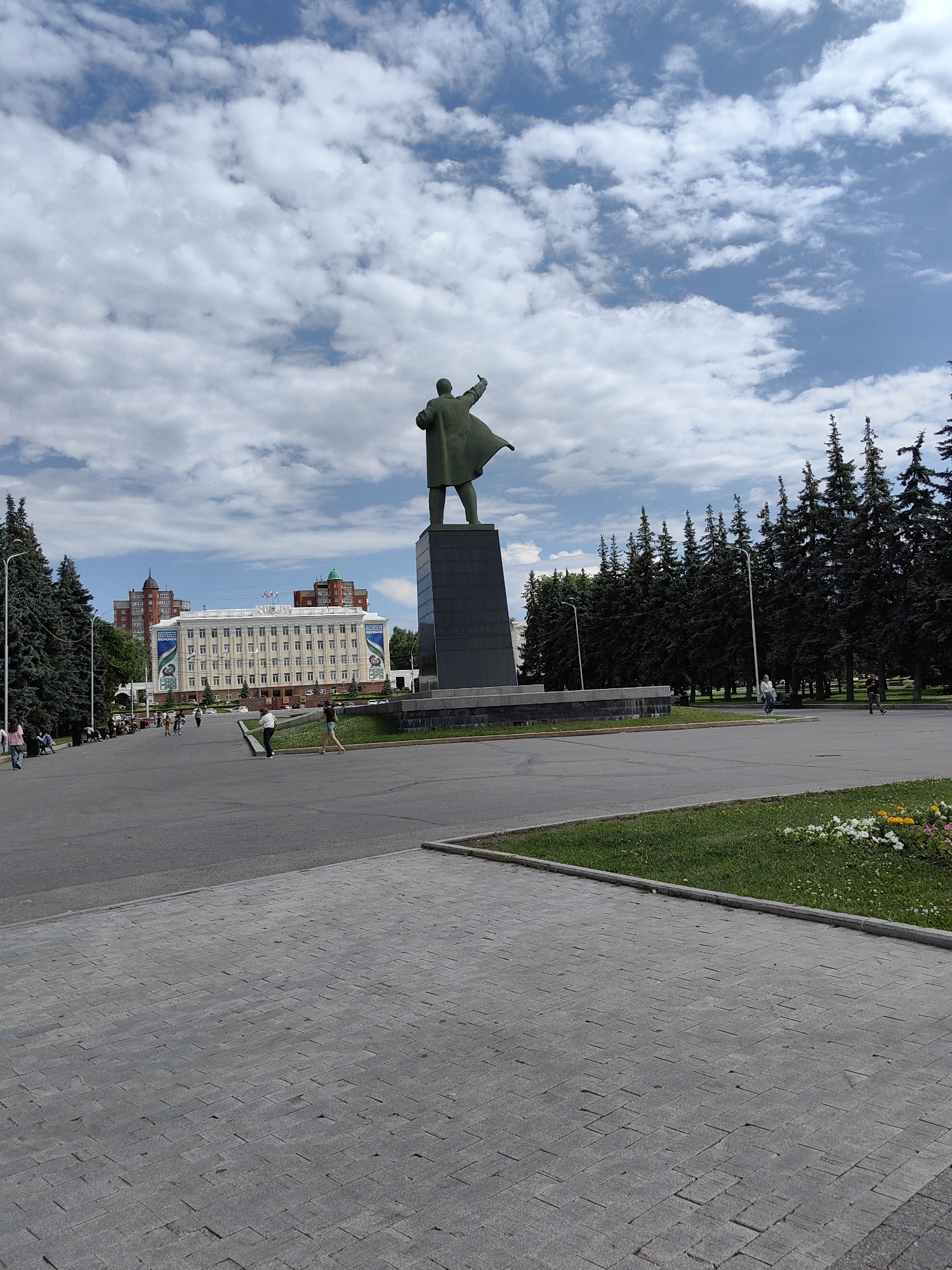 The image depicts a statue of Vladimir Lenin in Ufa, Russia, located in front of the Bashkir State Opera and Ballet Theatre. The statue, a prominent landmark, stands tall on a dark pedestal in a public square. The square is paved with grey stone tiles in the foreground and asphalt in the background. Several people are visible walking around the square, some seated on benches in the background. Tall evergreen trees line the edges of the square, and a large white building with red-trimmed windows, the opera house, is situated behind the statue. The sky above is partly cloudy with a mix of white cumulus clouds against a blue backdrop. The scene appears to be a sunny day in a well-maintained public space.