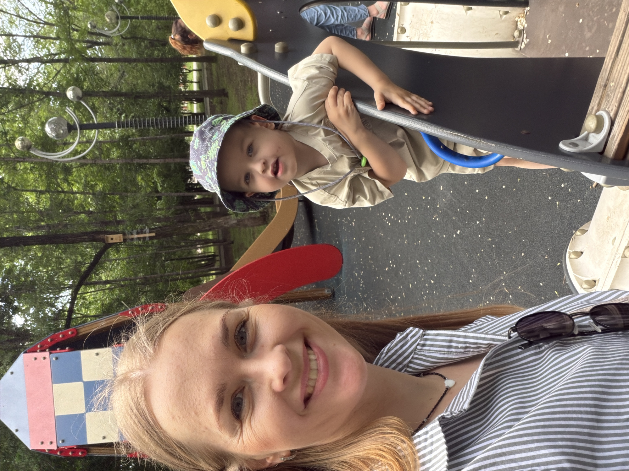 A selfie captures a blonde woman with blue eyes and a light smile, wearing a striped green and white shirt and a black beaded necklace with a white heart pendant. To her right, a young boy with light brown hair, wearing a tan short-sleeved shirt and a patterned bucket hat, is partially visible. He is holding onto a dark grey playground structure with a blue handle. The background reveals a park with trees, a colorful playground slide in the shape of a rocket, and lampposts. The ground is covered with dark grey safety surfacing and scattered leaves. A portion of another person's legs wearing blue jeans and sandals is visible on the right side of the frame. The scene appears to be a playground in Khabarovsk, Russia.