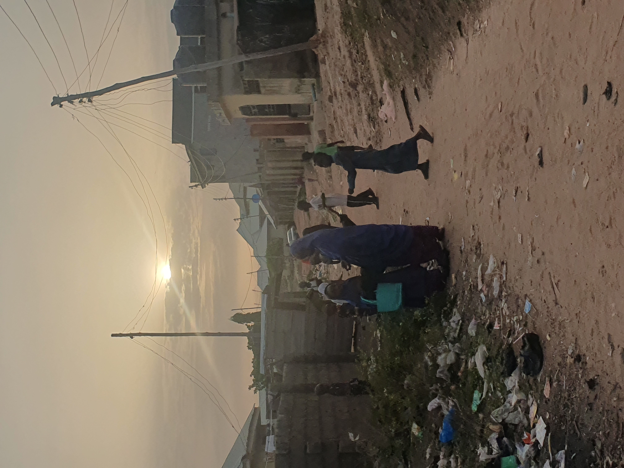 The image depicts a street scene in Lokoja, Nigeria, at what appears to be dusk or dawn. The sun is low in the sky, obscured by haze. Power lines are visible overhead. A dirt road dominates the scene, with litter scattered along its edge. Several people are present, including adults and children, some of whom are walking or running along the road. Buildings of simple construction line the street. The environment is dusty and shows signs of poverty.