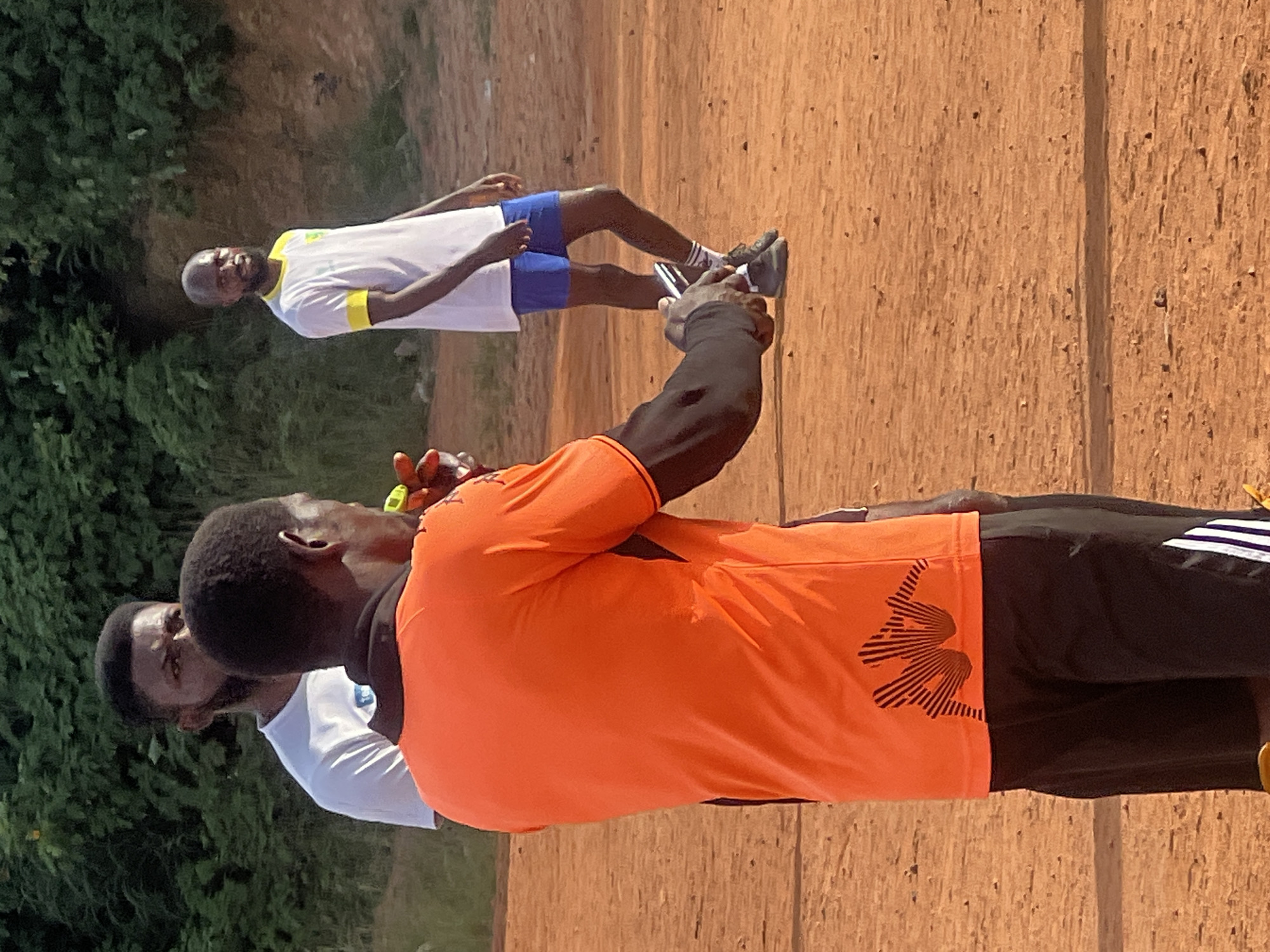 An outdoor, medium shot shows three men on a reddish-brown dirt field, possibly a sports pitch, with green foliage in the background. The man in the foreground, viewed from the back, wears an orange shirt with a black collar and black shorts with white stripes on the side. He is holding a mobile phone in his right hand, appearing to be recording or taking a picture. A second man, partially visible behind and to the left of the foreground man, wears a white t-shirt and has a beard. A third man stands in the mid-ground to the right, wearing a white t-shirt with yellow trim and blue shorts. The lighting suggests it is daytime, with the sun casting shadows. The overall scene depicts an informal outdoor gathering, likely related to sports or recreation, given the attire and environment. This scene could be located in a park or a less formal sports facility within the city of Jos, Nigeria.