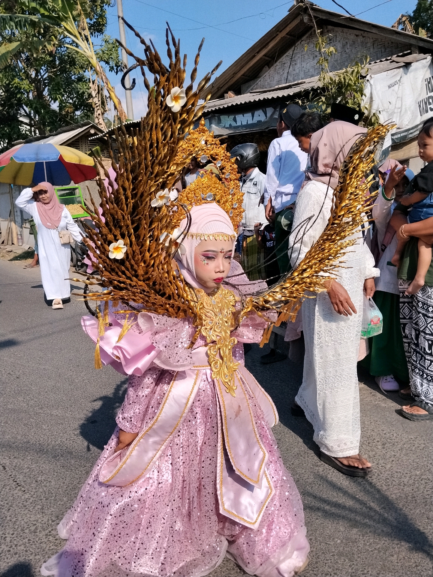 In Tlogosari, Indonesia, a young girl in a flamboyant pink costume with a sequined dress and elaborate gold headdress is the central focus of this image. She is adorned with large, golden wing-like structures made of dried branches, accented with white flowers. Her face is painted with dramatic makeup, including colorful eyeshadow and red lipstick. She is walking on a paved road, casting a shadow. To her left, a woman in a white dress and pink hijab is standing under a colorful umbrella, shielding her eyes from the sun. Behind the girl, several people are walking, including a woman in a white lacy dress, a child being held, and other individuals wearing traditional Indonesian attire. The background shows a street scene with buildings, some with corrugated metal roofs and bamboo walls, suggesting a rural or semi-urban setting. The sky is clear and blue.