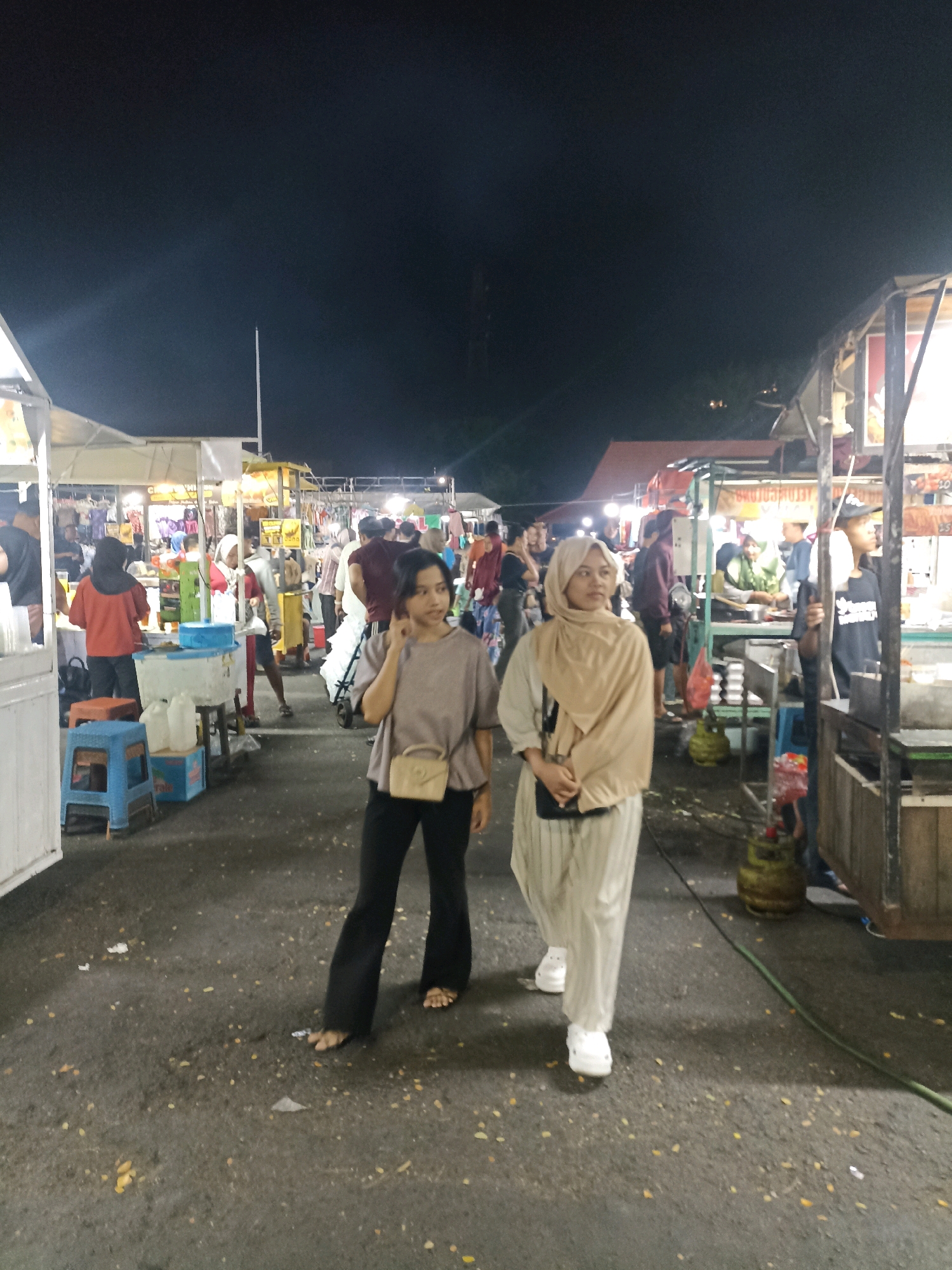 Two young women walk through a bustling night market in Surabaya, Indonesia. The woman on the left, with short dark hair, wears a light brown t-shirt, black flared pants, and a small beige purse across her chest. The woman on the right wears a beige hijab, a light-colored tunic over striped pants, and white clogs. They are walking away from the camera, on a paved ground littered with fallen leaves. In the background, numerous market stalls are illuminated by bright lights, displaying various goods and food items. People mill about the market, some browsing, others attending to their stalls. A white tent structure is visible on the far left, and a gas cylinder is present near a stall on the right. The overall scene depicts a lively evening at a street market.