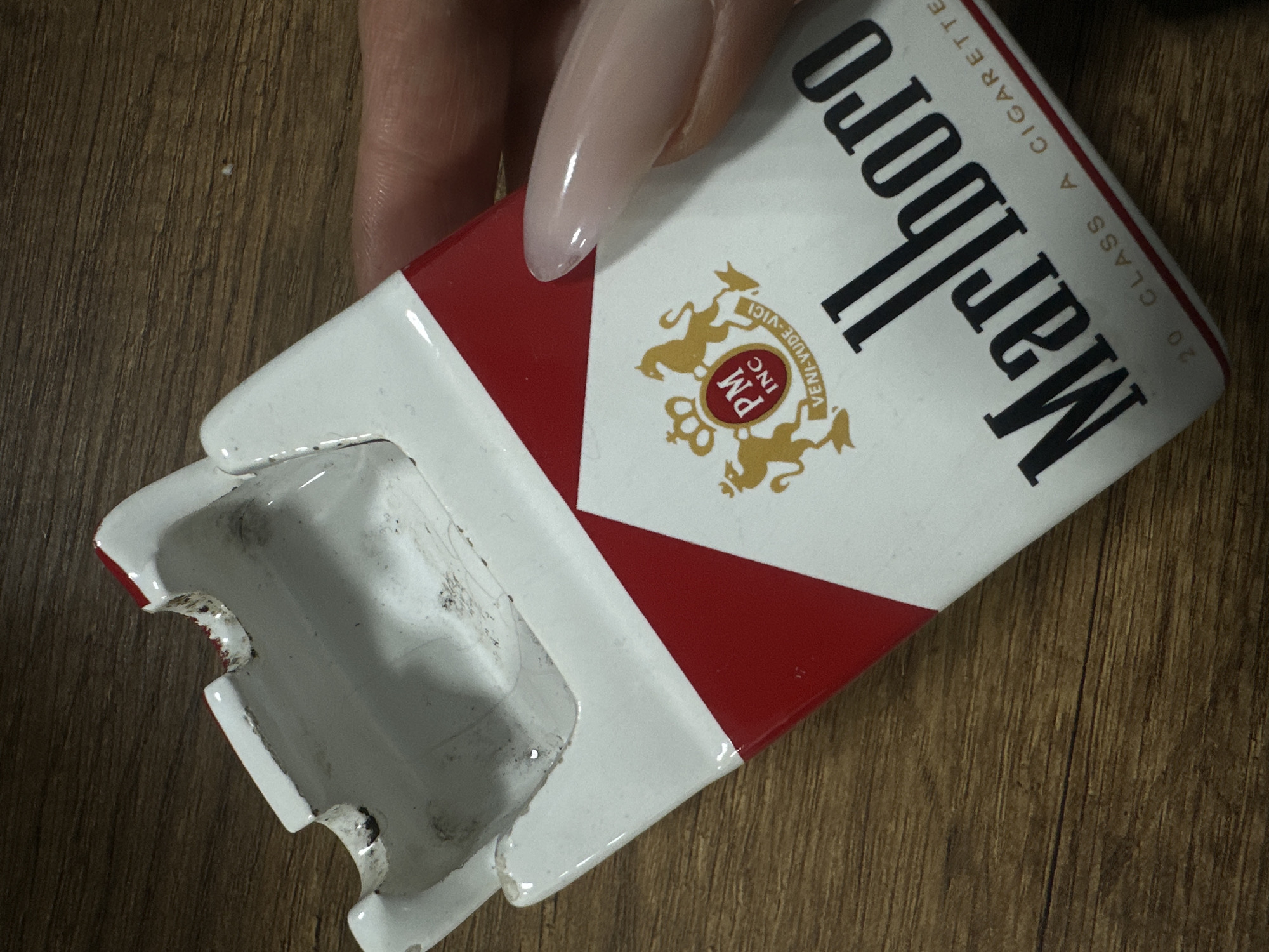 A close-up shot depicts an open, empty, enamel cigarette pack of Marlboro cigarettes held by a human hand with a long, manicured fingernail. The cigarette pack is positioned diagonally in the frame, with the open top angled toward the lower left. The Marlboro logo is visible, with the white and red color scheme and the brand name in black lettering. The pack is worn, with chipped enamel revealing the underlayer. The background is a wooden surface with a visible grain pattern. The image does not contain enough information to determine a location in Yerevan, Armenia.