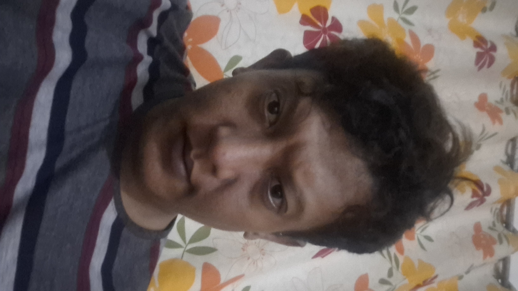 A medium shot captures a selfie of a person's face and upper chest, with a floral-patterned curtain in the background. The person has dark, somewhat messy hair and dark eyes. They are looking directly at the camera with a neutral expression. The curtain behind them features large, stylized flowers in shades of yellow, orange, and red, set against a light background. The person is wearing a gray t-shirt with horizontal stripes of dark blue and maroon. The image appears to have been taken indoors. There is no information available to indicate the location in Cibinong Hilir, Indonesia.