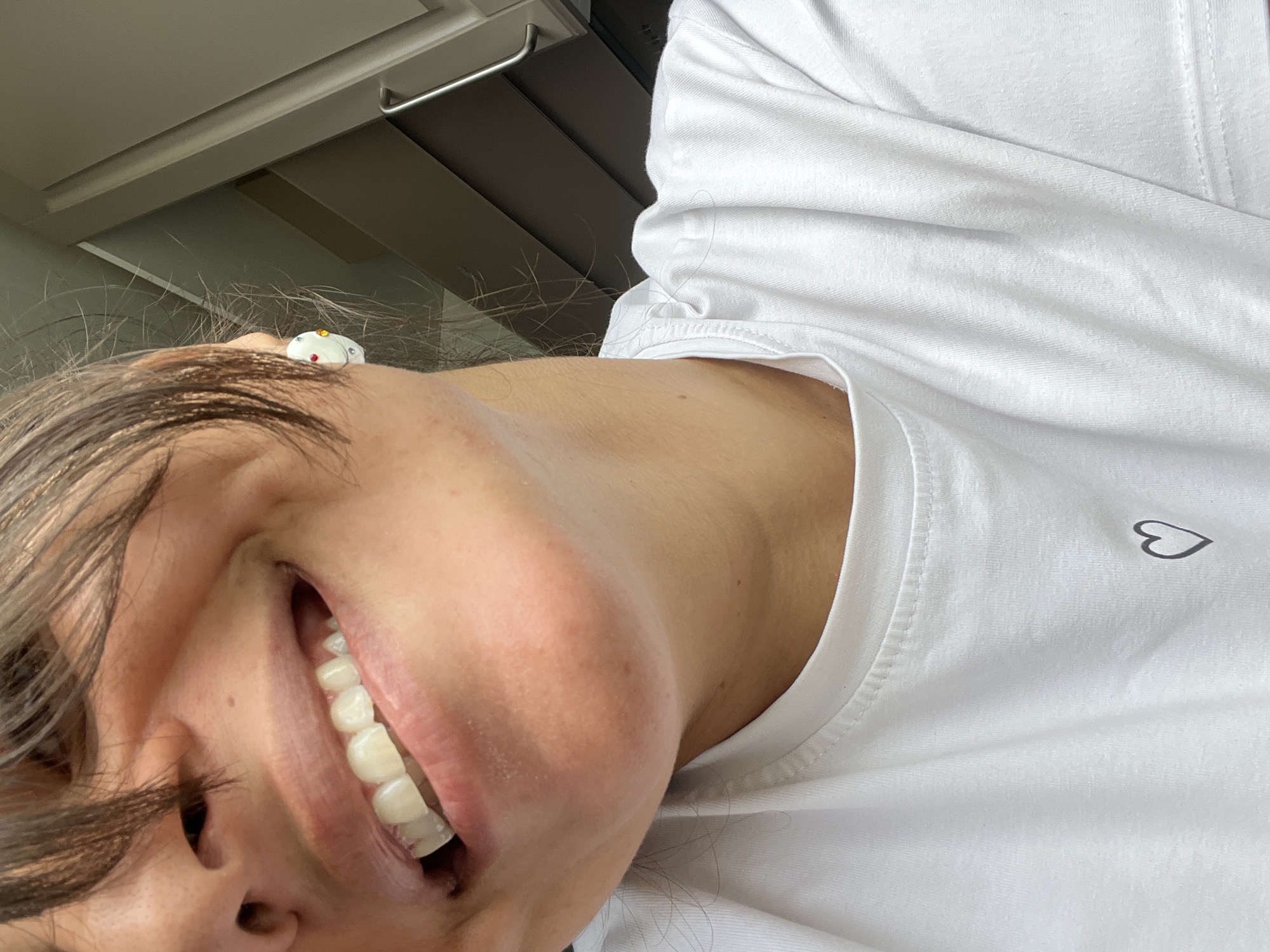 A close-up, waist-up shot captures a woman with dark hair and fair skin wearing a white t-shirt adorned with a small black heart graphic. She is smiling, revealing her teeth, and her right ear is visible with a decorative white earring featuring colorful rhinestones. The background consists of what appears to be kitchen cabinetry in muted tones of brown and beige, suggesting an indoor domestic setting. The lighting is soft, casting subtle shadows. There is no discernible indication of the location being Saint Petersburg, Russia.