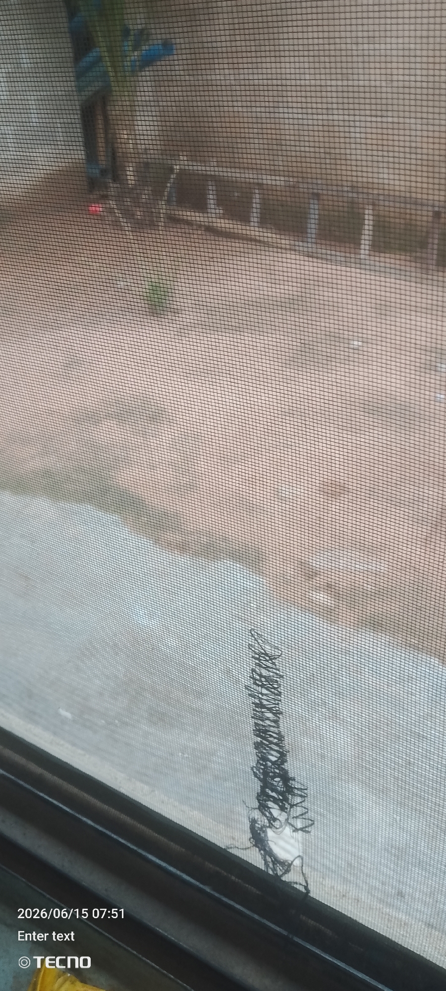 The image shows a view through a window screen. The screen has a tear with some black threads hanging from it. In the background, there is a sandy or dirt ground. There are some objects that appear to be a ladder and possibly some vehicles, but they are blurred and indistinct due to the screen and distance. In the lower left corner, there is a timestamp "2026/06/15 07:51" and the text "Enter text" and "TECNO".