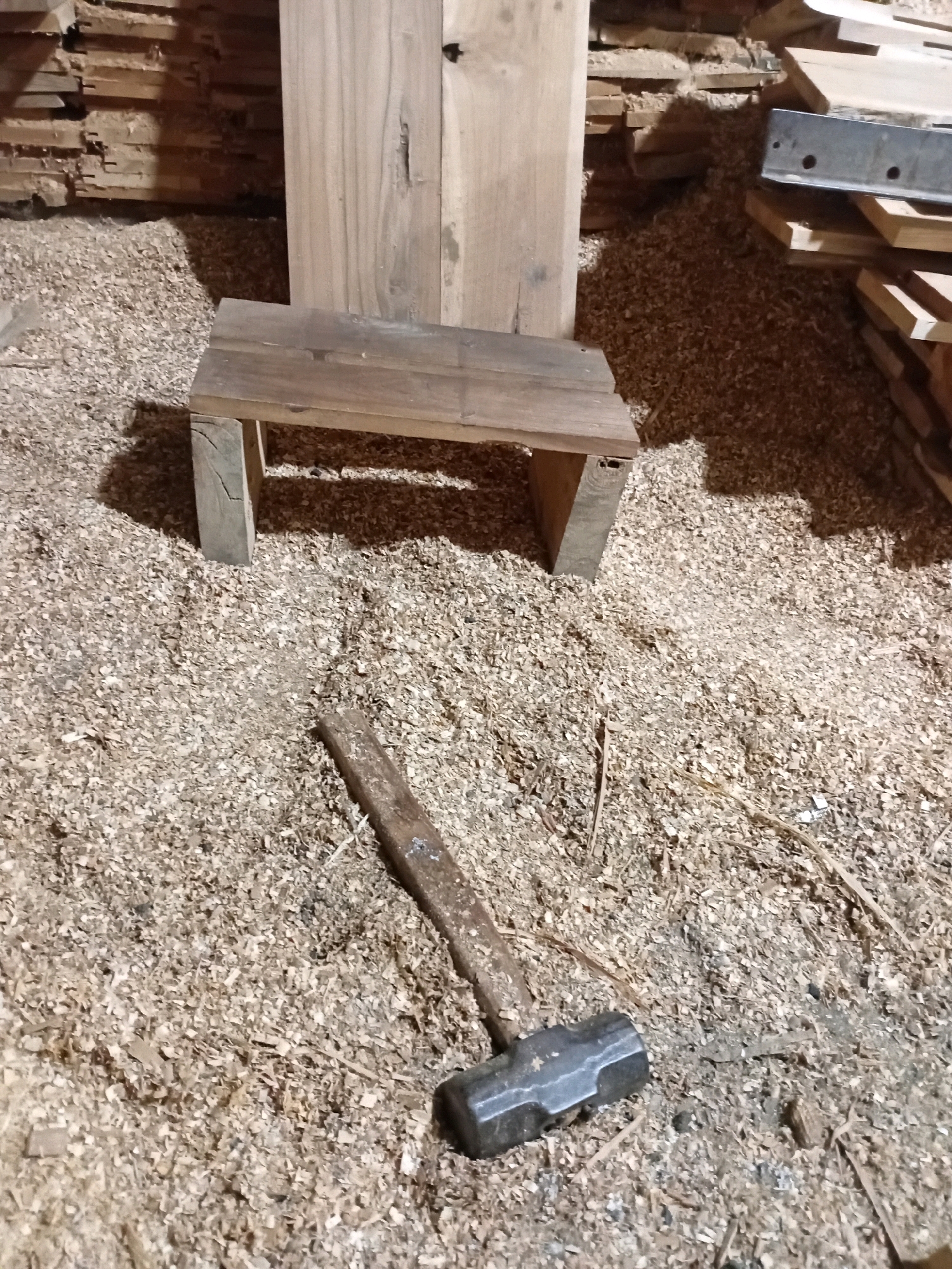 This image captures a close-up, low-angle view of a workshop floor covered in wood shavings. In the foreground, a metal sledgehammer lies horizontally on the shavings, with its head to the right. Behind it, a small, rustic wooden bench with two visible legs is positioned. The bench appears to be constructed from rough-hewn planks. The background shows stacks of lumber and larger wooden planks, suggesting a woodworking environment. The lighting is dim, casting shadows that add depth to the scene. The overall impression is one of a working woodshop. The location in Playen, Indonesia, is noted but not visually represented in the image itself.