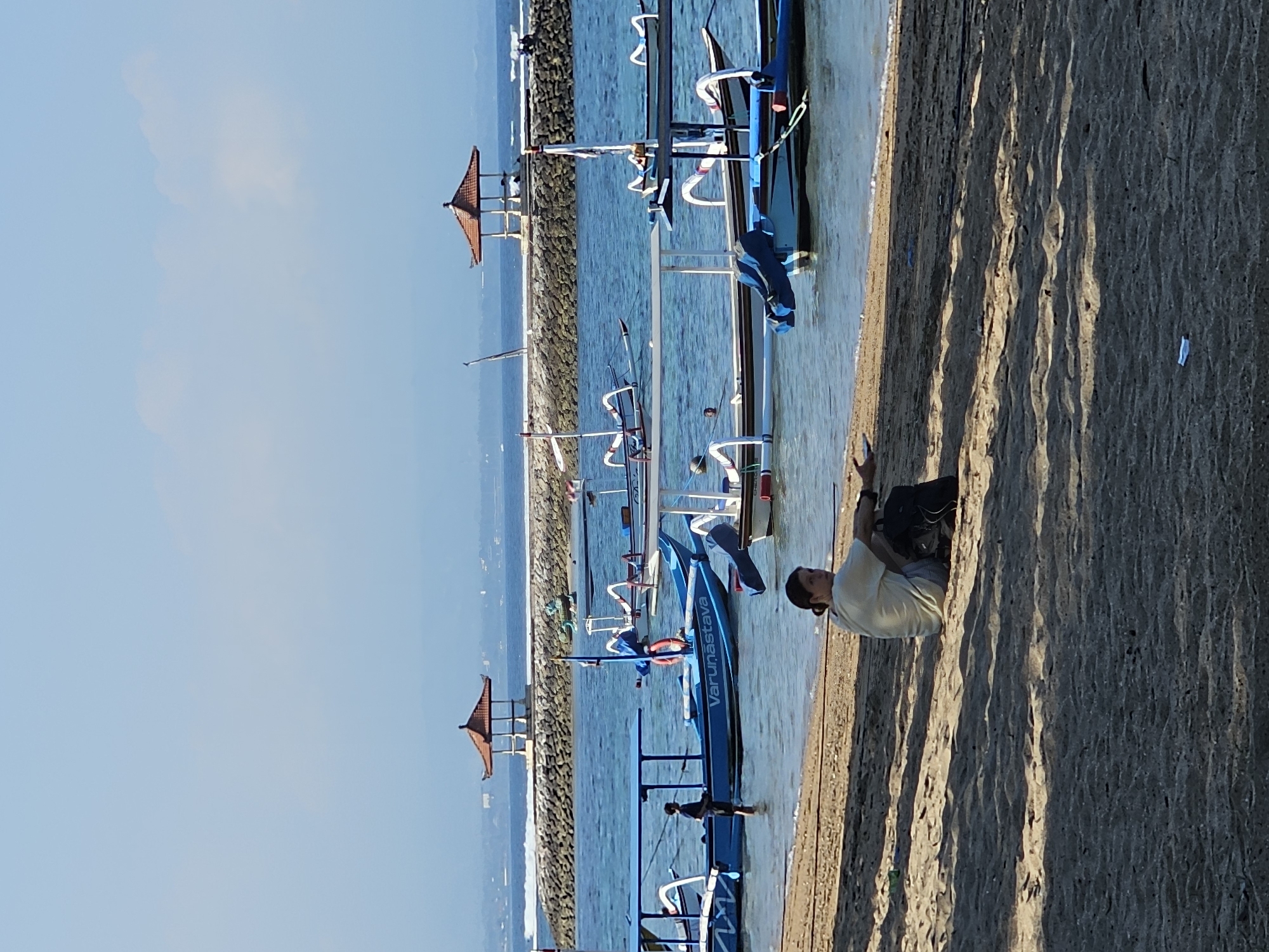The image depicts a tranquil beach scene in Denpasar, Indonesia, with several traditional Indonesian fishing boats, known as jukung, moored in the shallow, clear blue water. In the foreground, a person is seated on the sandy shore, facing away from the camera and towards the water. The individual is wearing a light-colored t-shirt and patterned shorts, with a black backpack placed beside them. Their posture suggests a moment of relaxation or contemplation.
In the middle ground, a man stands in the water beside a bright blue jukung, which has the name "Varunastav" written on its side. Other jukung boats, identifiable by their distinctive outriggers and colorful decorations, are also visible, some with awnings providing shade. The water is calm, with gentle ripples on the surface.
In the background, a breakwater constructed from large stones separates the calmer waters of the bay from the open sea. Two octagonal gazebos with reddish-brown tiled roofs, characteristic of Balinese architecture, are positioned on the breakwater, offering vantage points overlooking the ocean. Beyond the breakwater, the horizon is marked by a hazy outline of land and the vast expanse of the sky, which transitions from a pale blue near the horizon to a deeper shade of blue overhead, with a few wispy clouds. The sunlight casts distinct shadows on the sand, indicating it is likely late afternoon or early morning. The overall atmosphere is peaceful and serene, typical of a tropical coastal environment.