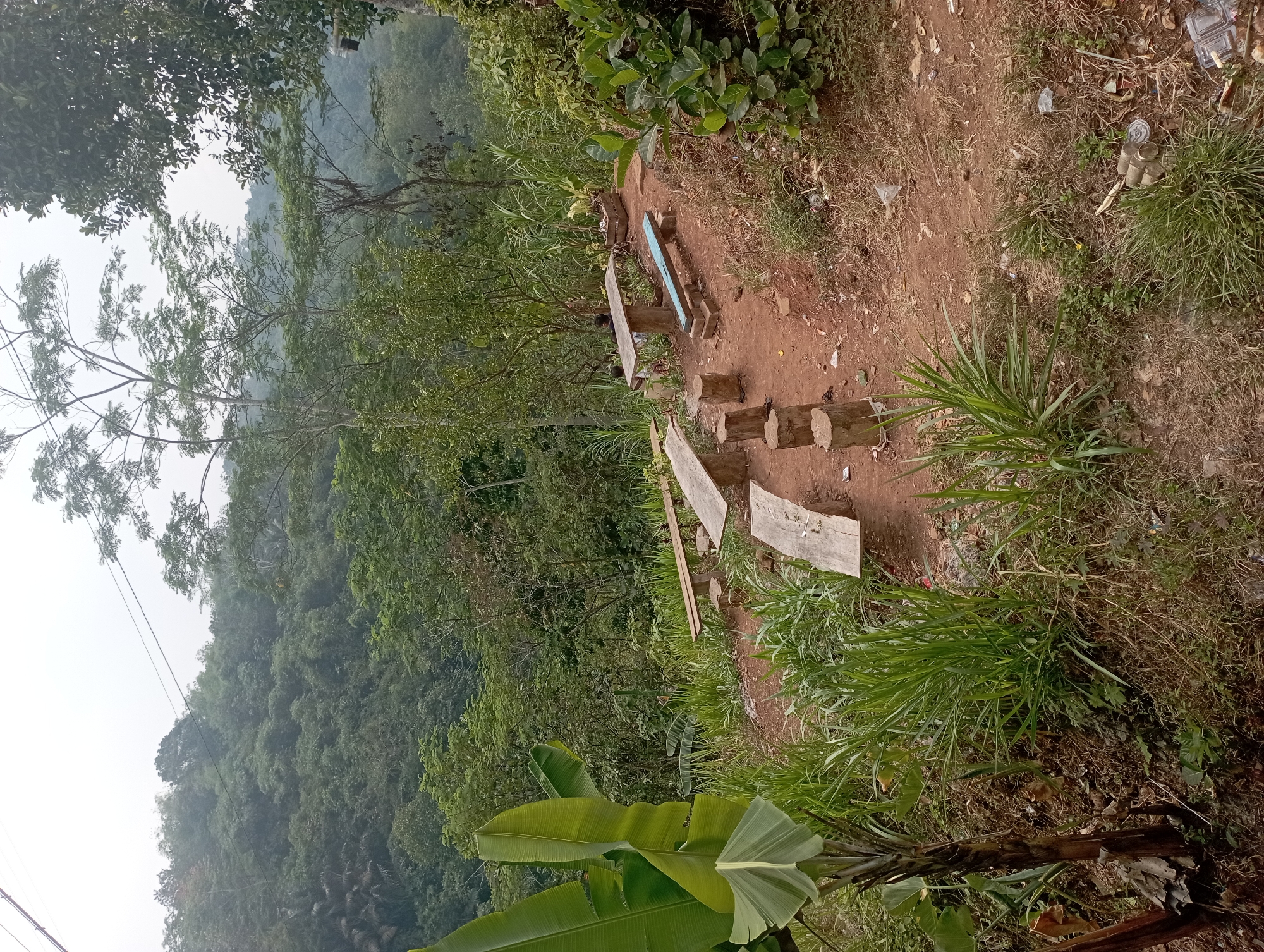 The image depicts an outdoor seating area on a hillside, likely in Manonjaya, Indonesia, overlooking a lush, forested valley. The foreground features a dirt path littered with some trash, including plastic bottles and cans. Several rustic wooden benches and log seats are arranged around a low table made of concrete and wood. Tall grasses and banana trees with large leaves are prominent on the left side of the frame. In the background, dense green foliage covers the undulating terrain, with a slightly hazy sky above. Power lines stretch across the upper portion of the image, disappearing into the trees. The scene appears to be a natural resting spot or a small, informal eatery with a view. There are no people visible in the immediate vicinity of the seating area, although faint figures can be seen in the distance near the concrete table.