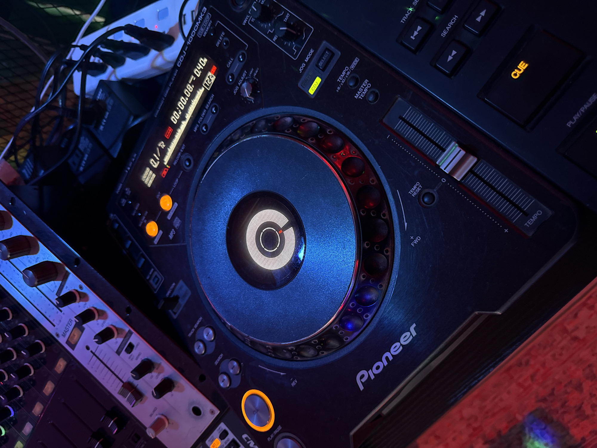 The image shows a close-up view of DJ equipment, specifically a Pioneer CDJ-1000MK3 and a mixer. The CDJ unit, a staple in DJ setups, is in the foreground, with its jog wheel prominently displayed and illuminated by blue and red lights. The digital display on the CDJ shows "00:00.08~040" and "122", suggesting a track is playing or cued. The mixer, positioned to the left, has several faders and knobs for controlling audio levels and effects.
The scene is set in what appears to be a dimly lit indoor environment, likely a bar, club, or DJ booth. The lighting is dramatic, with strong blue and red hues casting shadows and highlighting the equipment. Visible text on the equipment includes "Pioneer", "CDJ-1000MK3", "MP3 FOLDER SEARCH", "CUE", and "PLAY/PAUSE" on the CDJ, and "MASTER" on the mixer. The red and blue lighting, along with the close-up perspective on the professional DJ gear, suggest a setting where music is being played or prepared for performance, possibly late at night. The overall mood conveyed is one of active music production or performance. There are no people visible in the frame. The location context provided is Si Sunthon, Thailand.