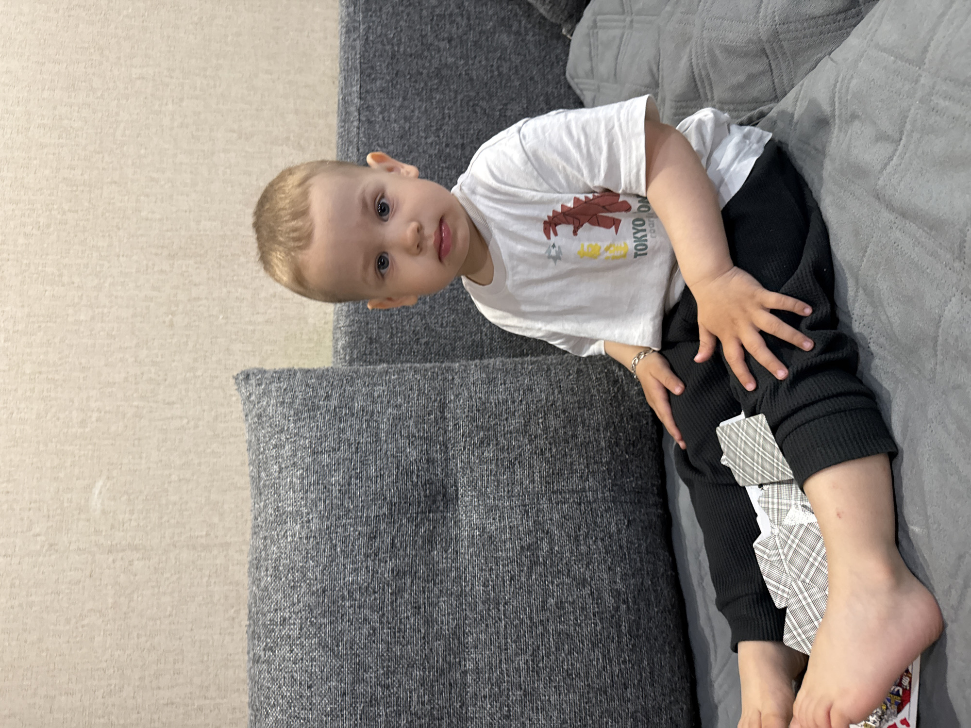 A child lies on a gray couch with a deck of playing cards positioned on their leg. The child is wearing a white t-shirt with a drawing of a dinosaur and black pants. The background appears to be a domestic interior setting. There is no indication of location in the city of Rusanovo, Russia.
