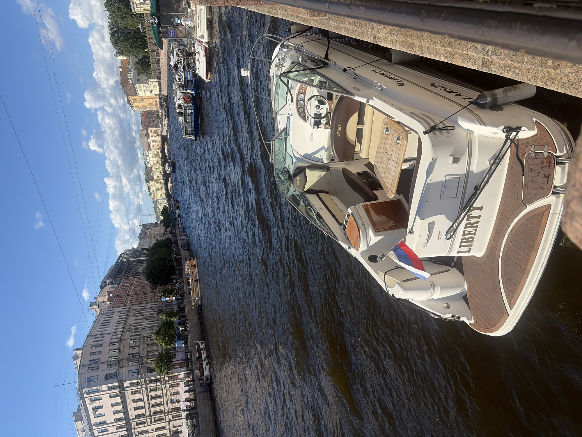 The image captures a sunny day on a river in Saint Petersburg, Russia, likely the Griboyedov Canal, judging by the distinctive architecture and canal boat traffic. In the foreground, a white Larson motorboat named "LIBERTY" is moored to a stone embankment. The boat features cream-colored upholstery, a wooden dining table, and a Russian flag displayed at the stern. The backdrop is lined with classical European-style buildings, typical of Saint Petersburg, with ornate facades and windows. Several other boats, including tour boats with passengers, are visible further down the canal, navigating the dark, rippling water under a bright blue sky dotted with white clouds. Overhead, power lines crisscross the sky. The scene conveys a sense of leisurely urban exploration and sightseeing on a pleasant summer day.
