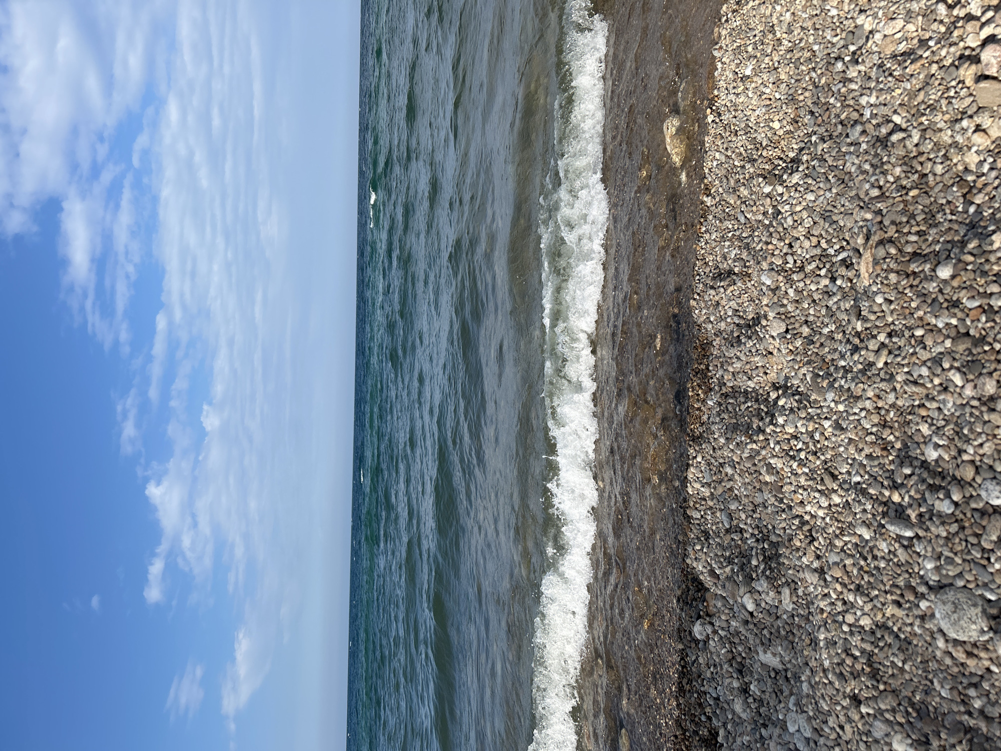 The image depicts a clear day at a pebble beach on the shore of Lake Baikal, near the village of Tolbazikha, Russia. The sky is a vibrant blue, with scattered white clouds. The water of Lake Baikal is a deep, dark teal color, with small waves lapping at the shore. The waves are breaking, creating white foam as they reach the pebbles. The beach itself is covered in smooth, grey and brown pebbles of various sizes. The overall impression is one of natural beauty and tranquility.