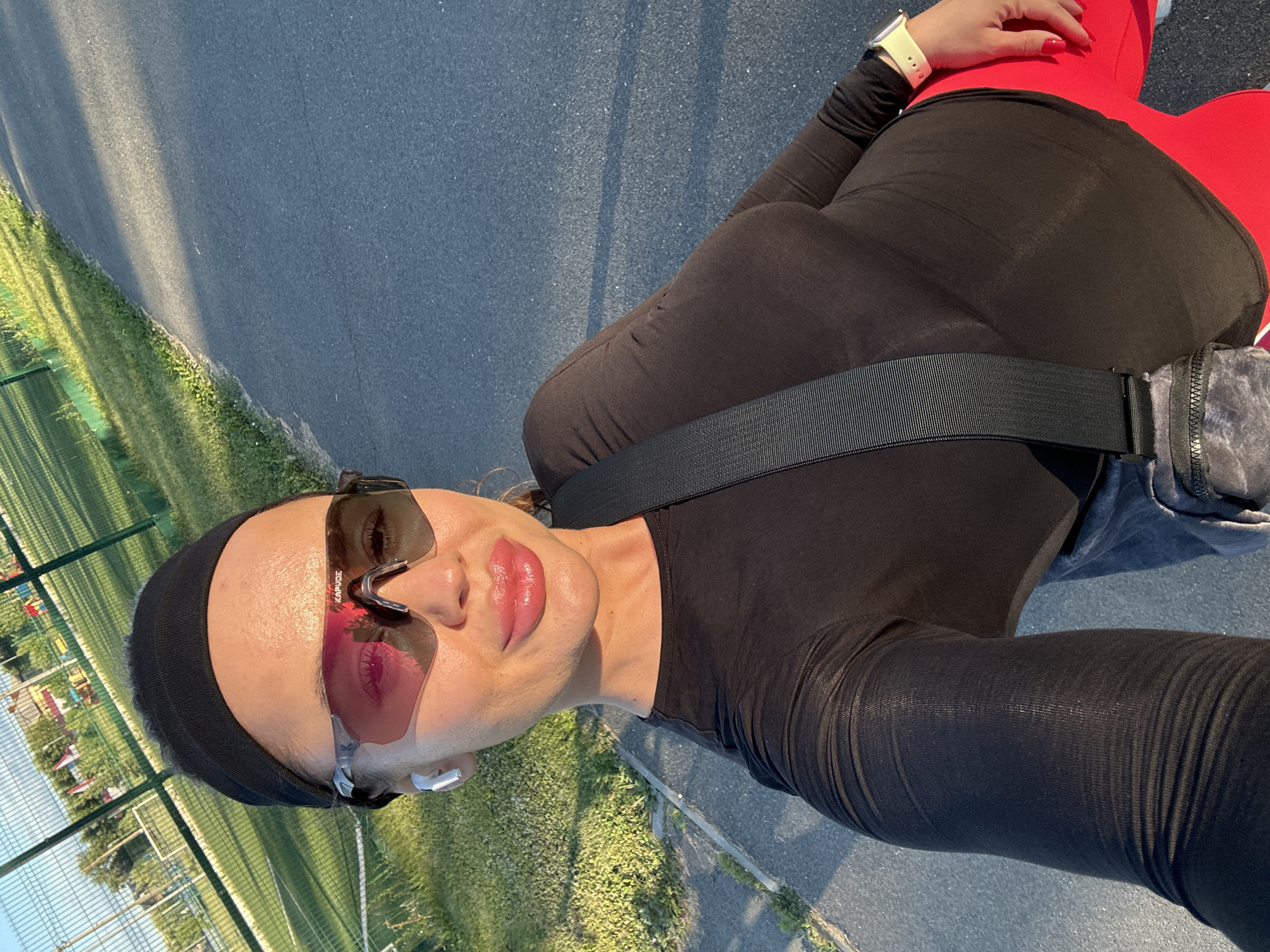 A woman is taking a selfie outdoors in Yuzhnouralsk, Russia. She is wearing a black long-sleeved shirt, red leggings, a black headband, and sunglasses. She has on white wireless earbuds and a smartwatch on her left wrist. A black strap crosses her chest from her right shoulder to her left hip. The background shows a grassy area with a fence, a sports field, and some buildings in the distance. The ground is asphalt. The lighting suggests it is late afternoon or early evening.
