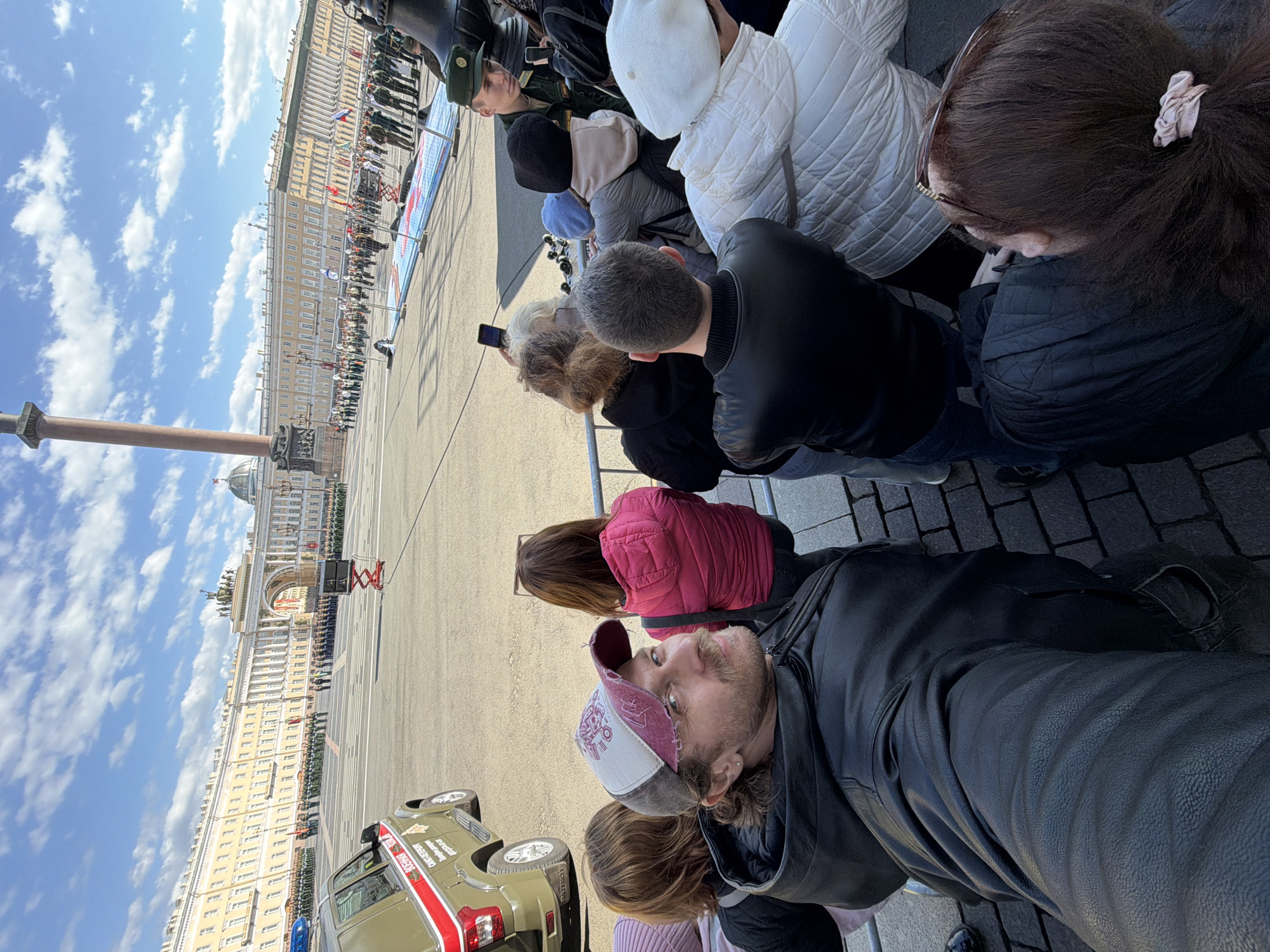 A street-level photograph depicts a crowd of people, seemingly observing an event in Saint Petersburg, Russia. The Alexander Column and the Winter Palace are visible in the background, indicating the scene is set on Palace Square. A military parade is underway. People are dressed in casual attire; several have their backs turned to the camera, while one individual is facing the camera, possibly taking the picture. A military vehicle is visible on the left side of the frame. The sky is partly cloudy.