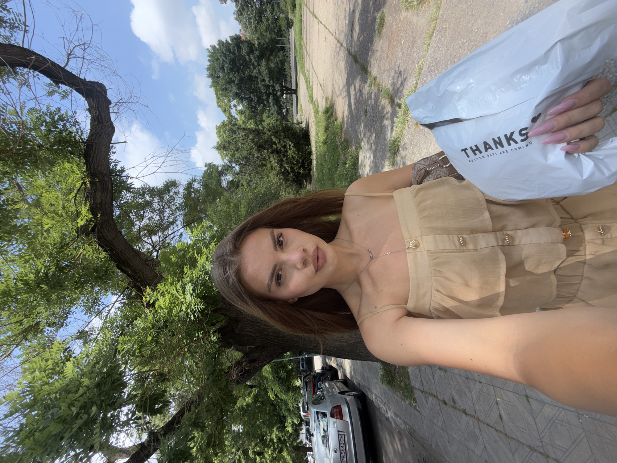 A young woman is captured in a selfie taken outdoors in Plovdiv, Bulgaria, under bright daylight. She is the central subject, looking directly at the camera with a neutral expression. She has long brown hair and is wearing a light-colored (beige or tan) strappy top or dress with button and ruffle details. A delicate silver-toned necklace with a small charm is visible around her neck.
In her right hand, she holds a white plastic bag or package, which has the word "THANKS" printed in black, accompanied by a small heart icon above it, and the phrase "BETTER DAYS ARE COMING" below. Her nails are long and painted a sparkly light pink. A portion of a patterned shoulder bag is also visible on her right side.
The background reveals an urban green space with lush, leafy trees framing the scene, particularly on the left. A clear blue sky with scattered white clouds is overhead, suggesting good weather. Below, a paved surface, likely a sidewalk or path, shows signs of wear with cracks and grass. Parked cars are visible on a street behind her, including a silver sedan with a Bulgarian license plate displaying "PB 5643 CH," confirming the Plovdiv location. The strong shadows indicate a sunny mid-day or afternoon setting.