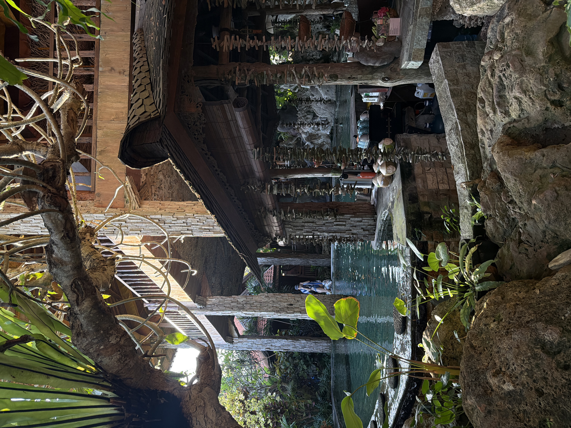 This media file captures an outdoor scene with a prominent water feature, likely a pool or stream, reflecting the surrounding architecture and vegetation. The architecture itself appears to be made of stone and wood, with exposed beams and a tiled roof. Hanging from the beams are numerous small objects, possibly decorative or commemorative. Large rocks and lush green plants frame the water, suggesting a naturalistic or tropical setting. In the distance, several people are visible near the water, some appearing to be seated. The overall impression is one of a tranquil, perhaps resort-like, environment. The lighting suggests it is daytime, with sunlight dappling through the foliage. The location cues, such as the tropical plants and the style of architecture, along with the provided location context of Mengwi, Indonesia, indicate a tropical or subtropical climate. The image is oriented vertically, with the scene appearing to flow from top to bottom as if viewed from above. There is some text visible on what appears to be a bottle in the background, reading "OATDRINK," and another small label that is illegible.