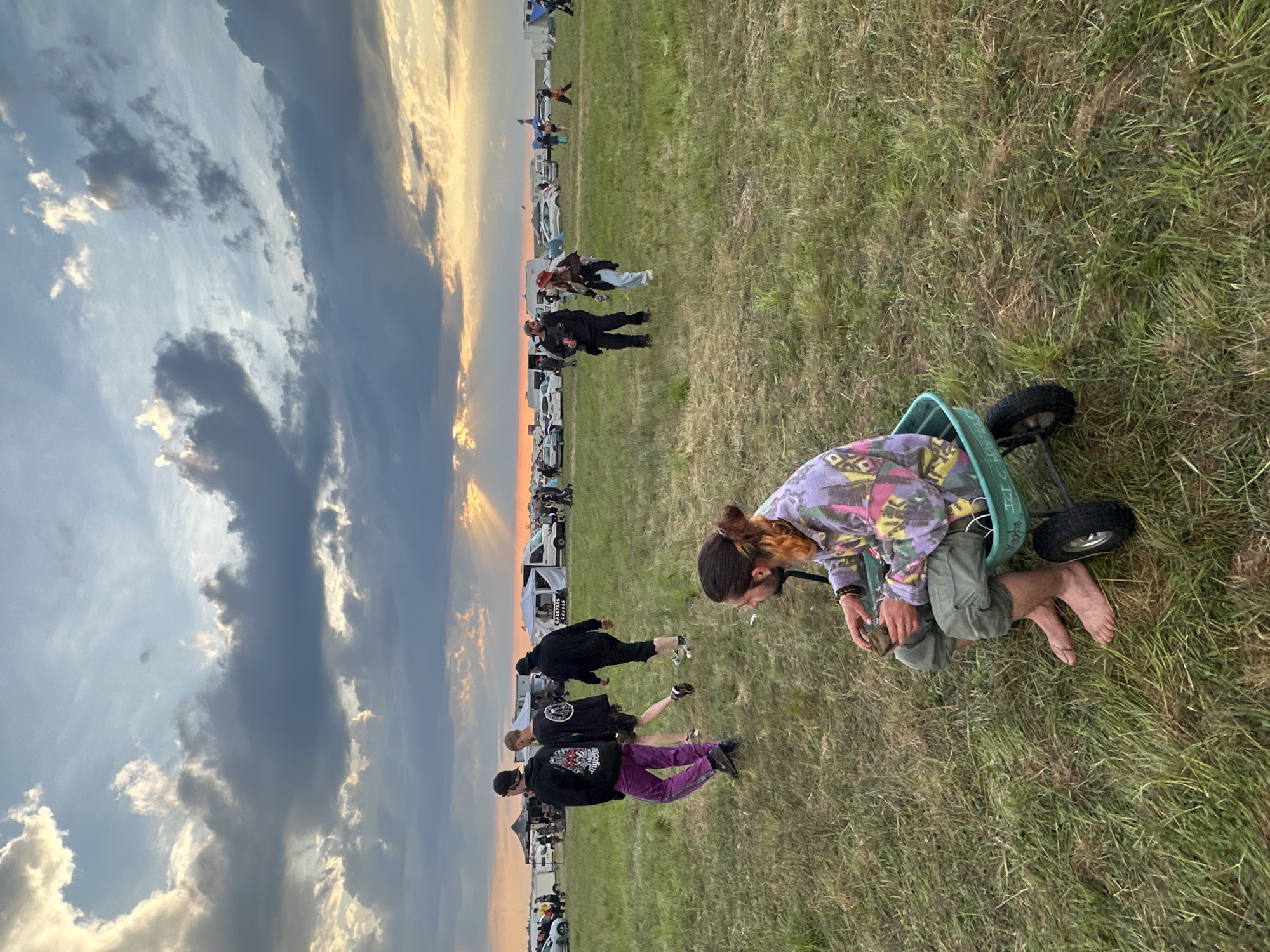 The image, taken outdoors in Bengy-sur-Craon, France, shows a grassy field setting at dusk. A young individual, seen from the side, is kneeling inside a small, wheeled, green cart. They are wearing a tie-dye top, green pants, and are barefoot. Their hair is pulled back. They appear to be examining or adjusting something in front of them. In the background, multiple individuals stand and walk in the same direction as the cart, with a line of parked vehicles visible further back. The sky in the background features a sunset with hues of orange and yellow. The lighting appears natural.